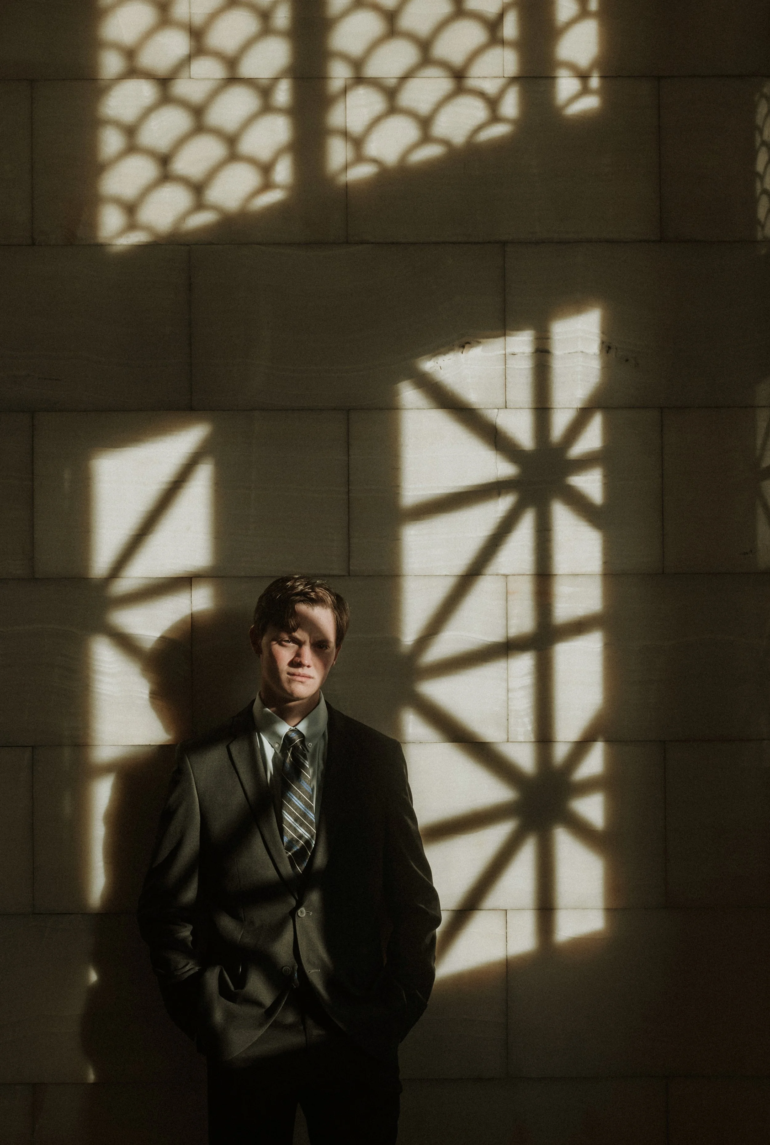 A young man in a suit and tie standing in front of a beige stone wall. Shadows of a balcony or railing and a large wheel or clock are cast on the wall behind him.