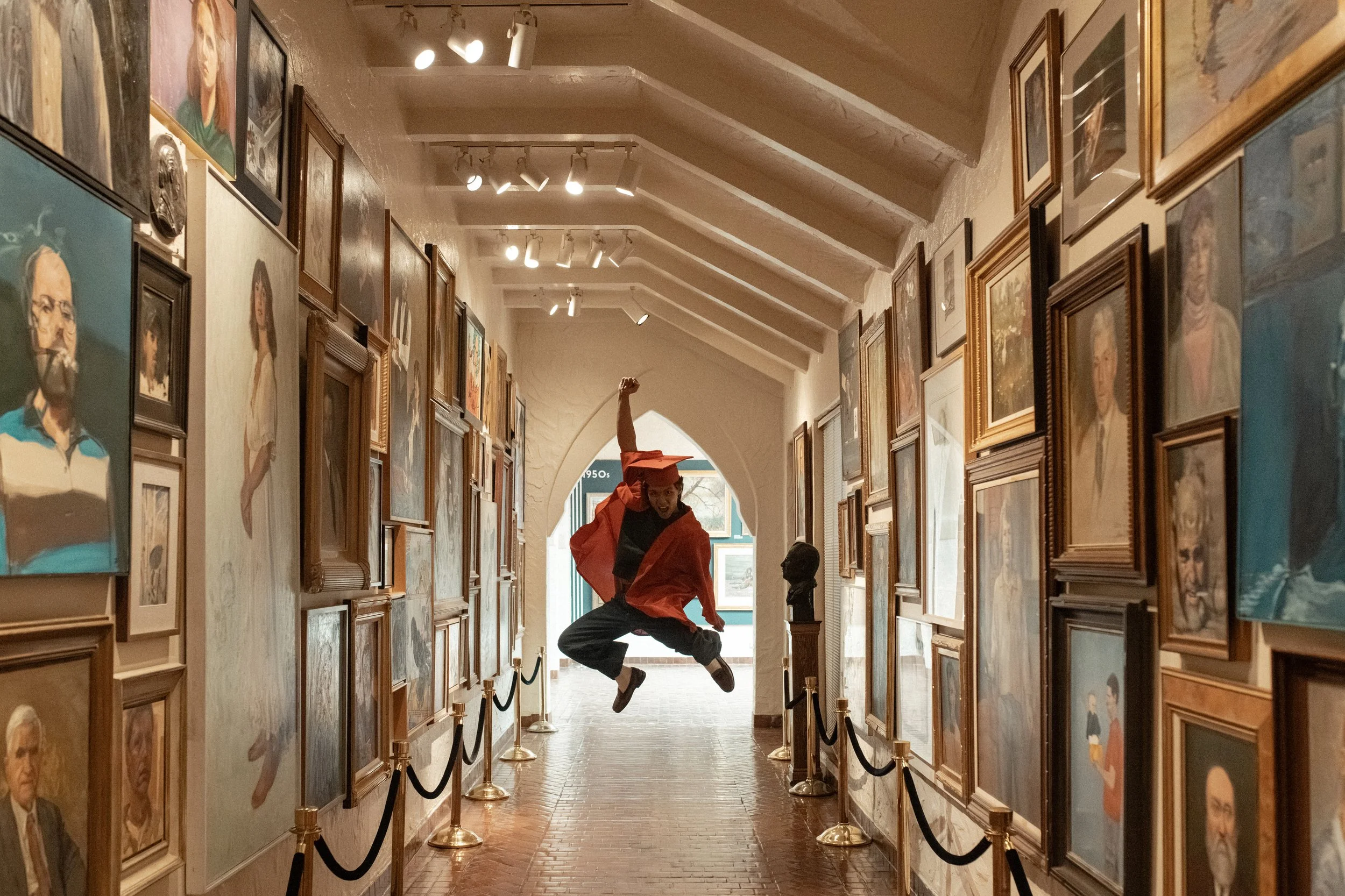 A person dressed in red and black is jumping in the air inside an art gallery with many framed paintings on the walls and a bust sculpture on a pedestal.