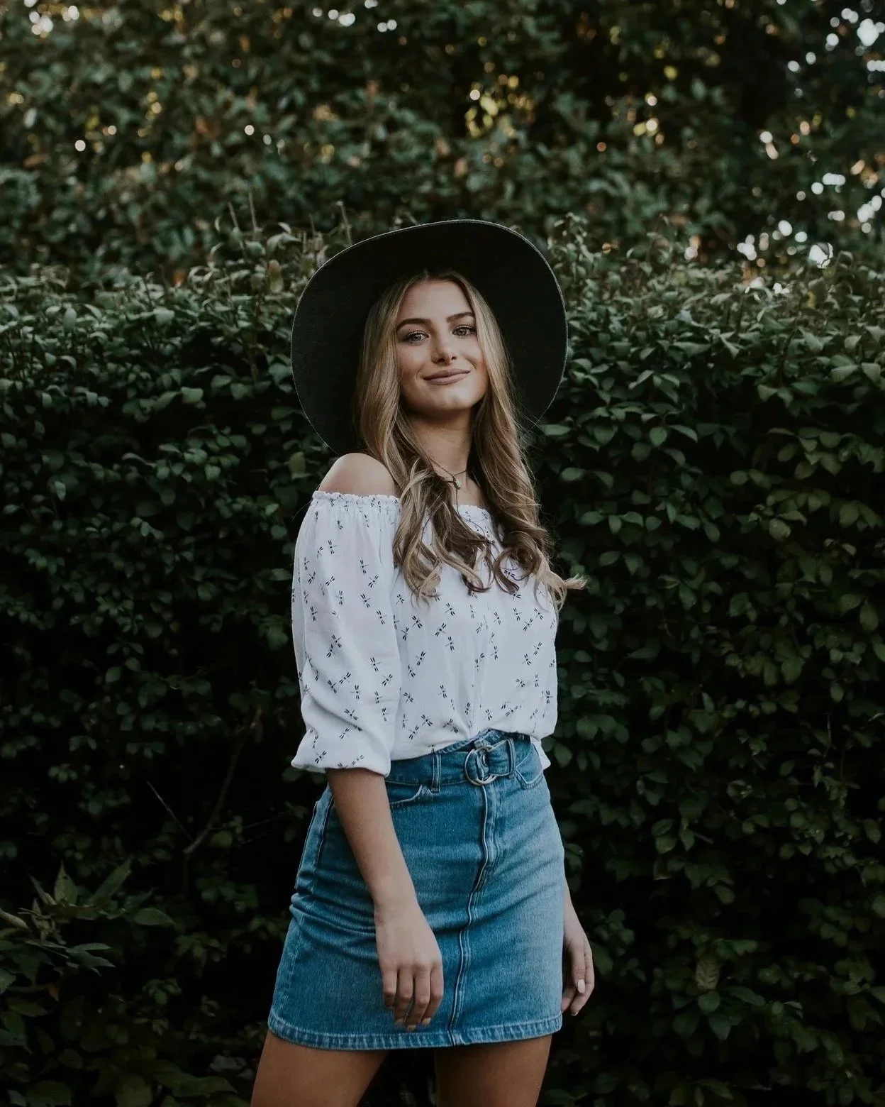 A young woman standing outdoors in front of green foliage, wearing a black wide-brimmed hat, a white off-shoulder blouse, and a denim skirt.