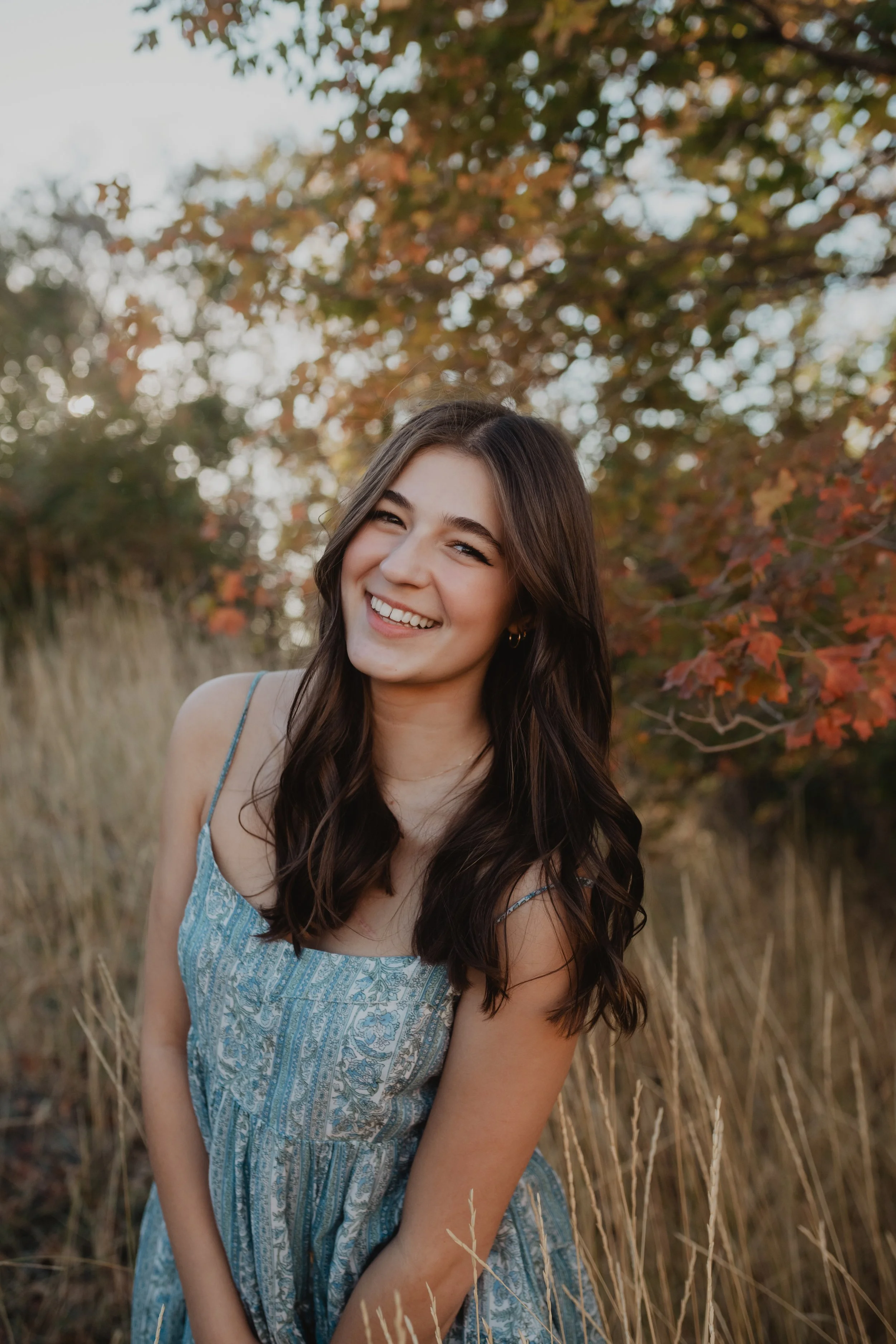Smiling young woman with long dark hair, wearing a light blue patterned dress, standing outdoors in a field of tall grass, with trees with autumn-colored leaves in the background.