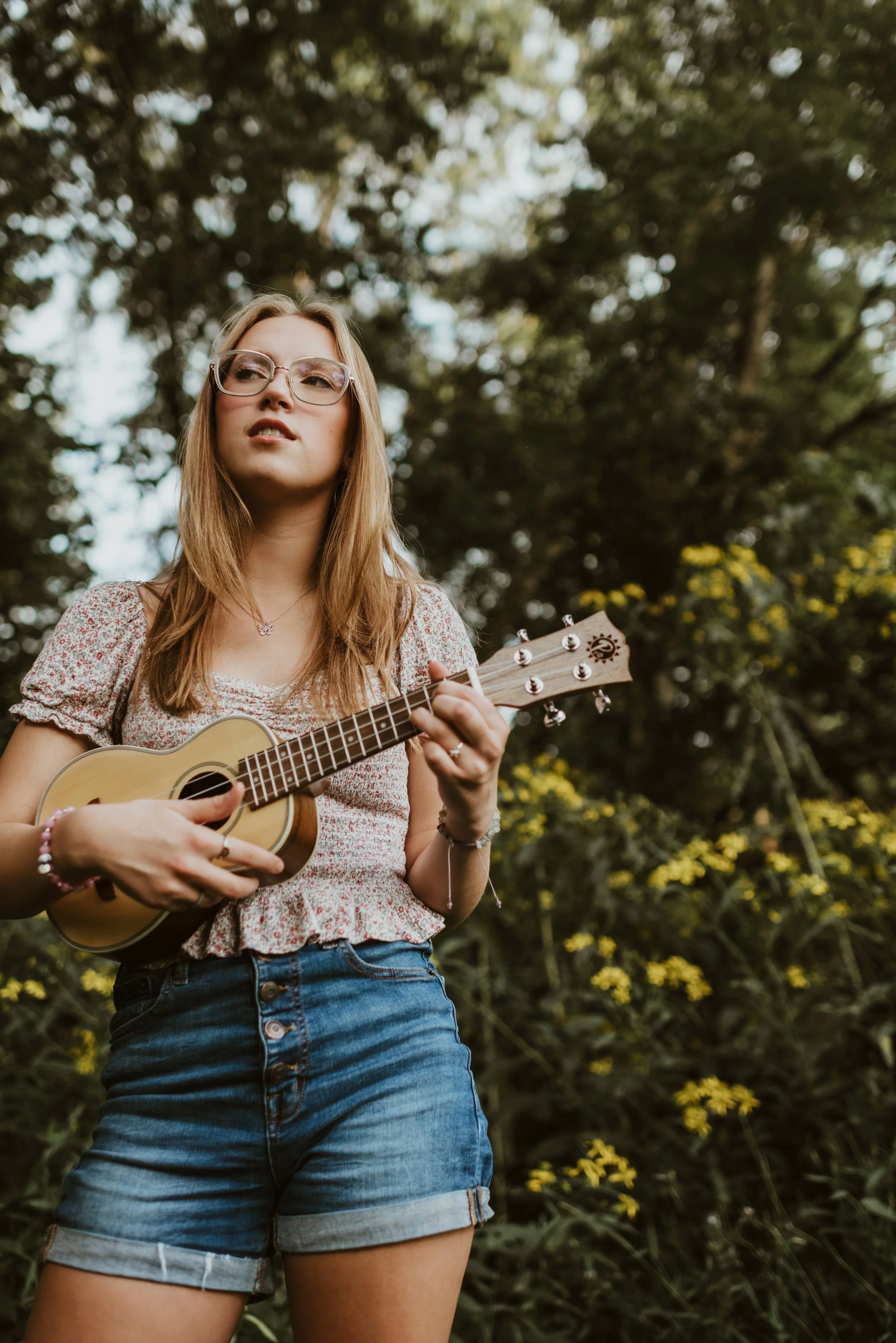 A young woman with long blonde hair, glasses, and a floral top playing a ukulele outdoors among green trees and yellow flowers.