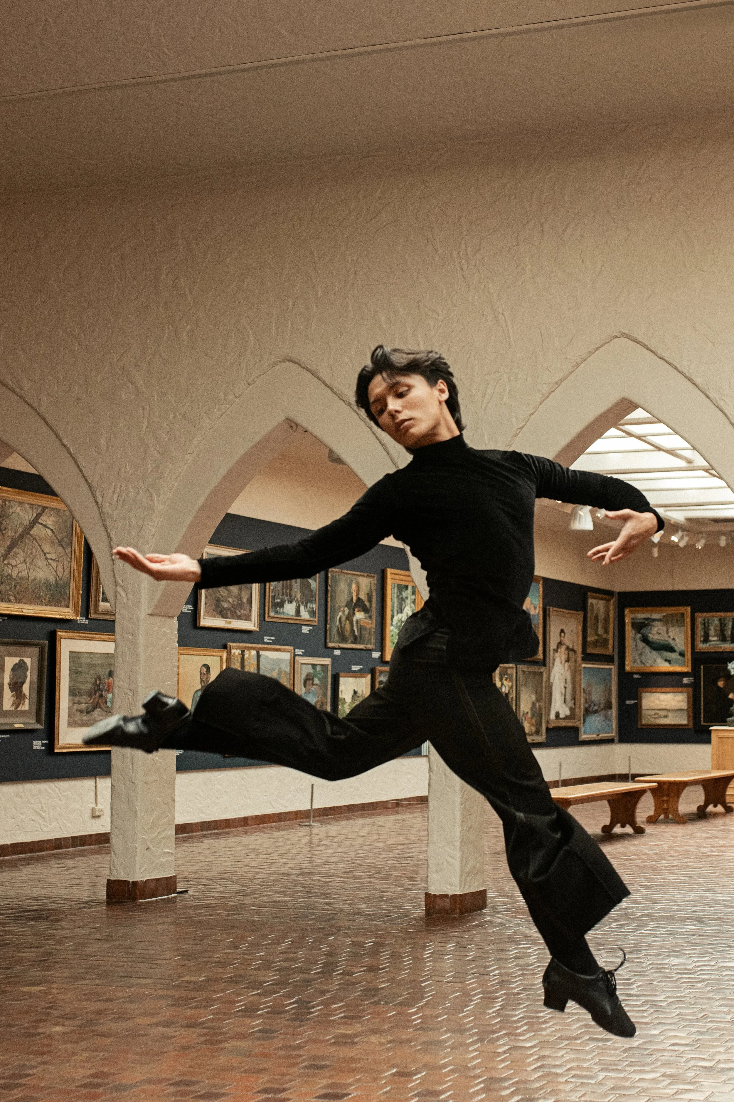 A young woman with short dark hair performing a dance leap in an art gallery. She is wearing a black turtleneck, black pants, and black shoes. The gallery has a curved ceiling and walls decorated with framed paintings.