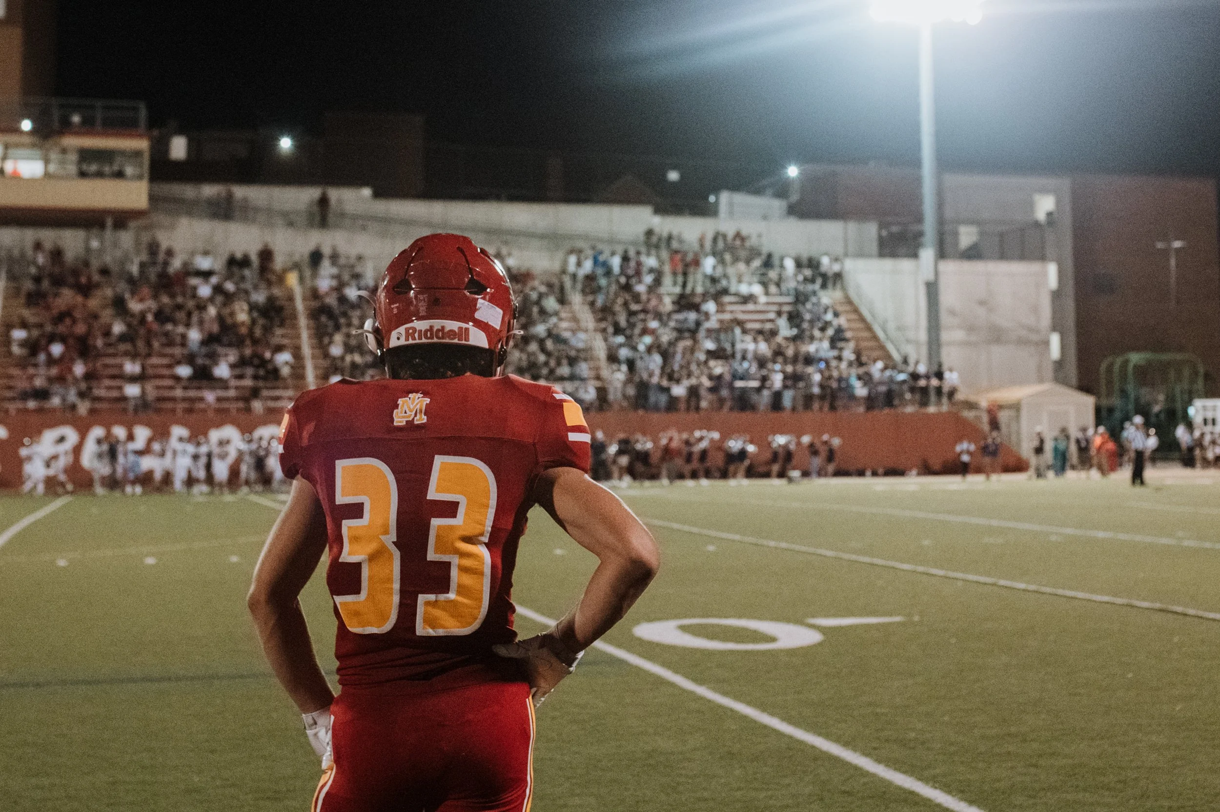 A football player wearing a red and yellow uniform with the number 33 on the back, standing on the field during a game at night with bright stadium lights, surrounded by spectators and other players.