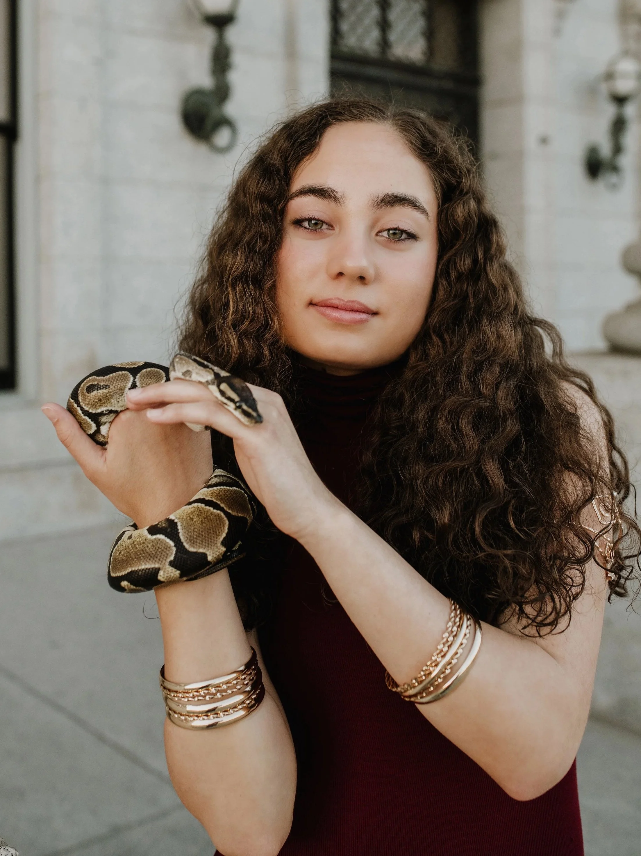Young woman with long curly hair holding a snake on her arm, standing outdoors near a stone building.