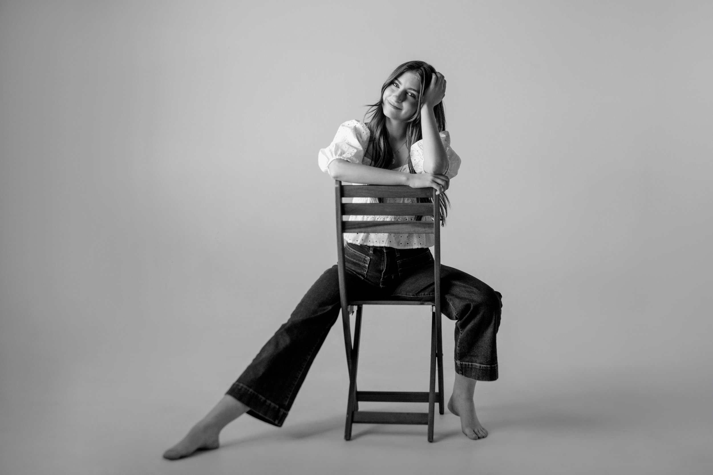 A black-and-white photo of a woman with long hair sitting on a chair, leaning on it, smiling, with one hand in her hair. Her other arm rests on the back of the chair, and one leg is extended while the other is bent.