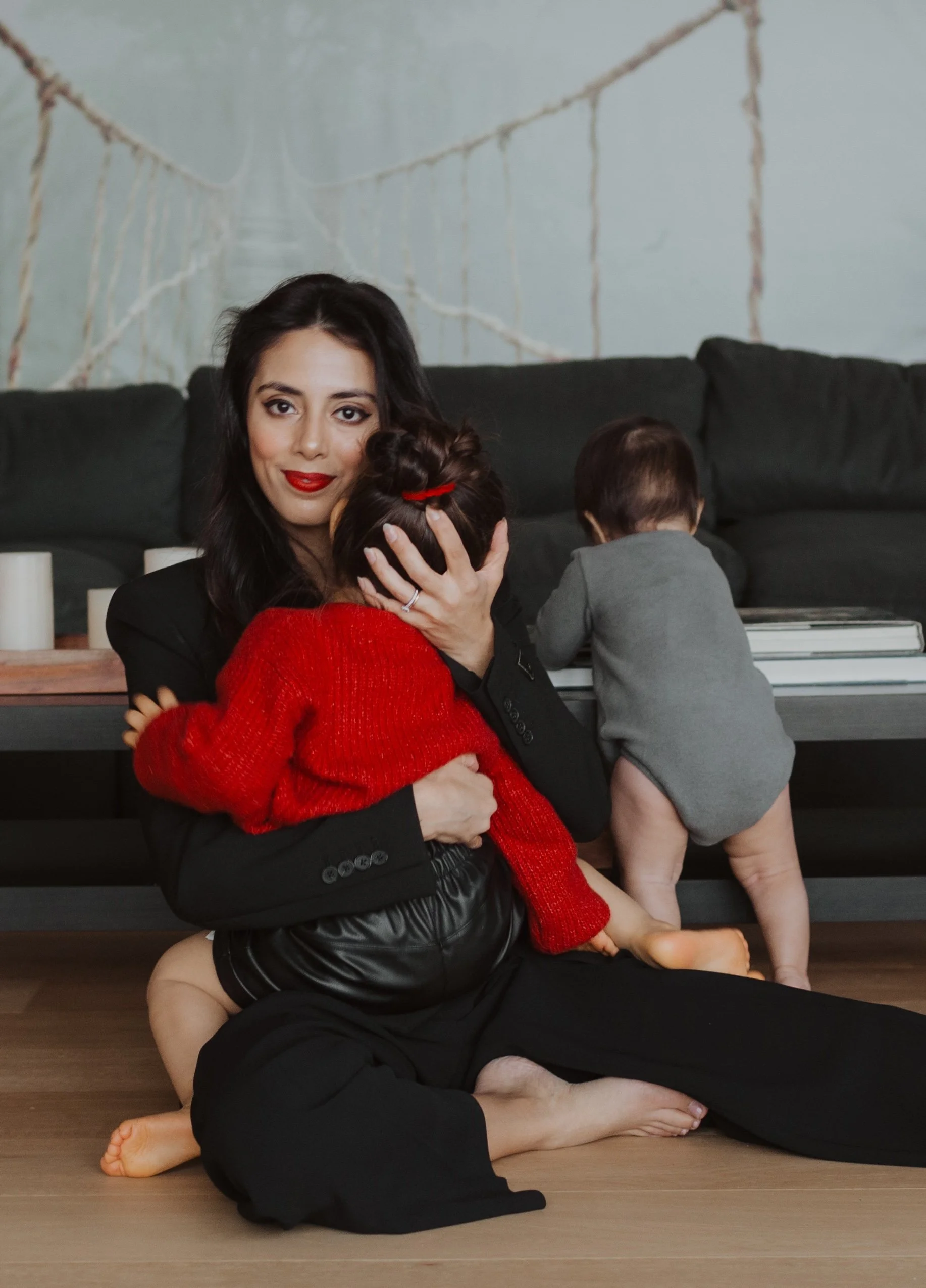 Basma dressed in black holding her daughter dark hair and red sweater, and son with dark hair in gray shorts, sitting on the floor with a black sofa and a light blue wall with a mural of a bridge in the background.