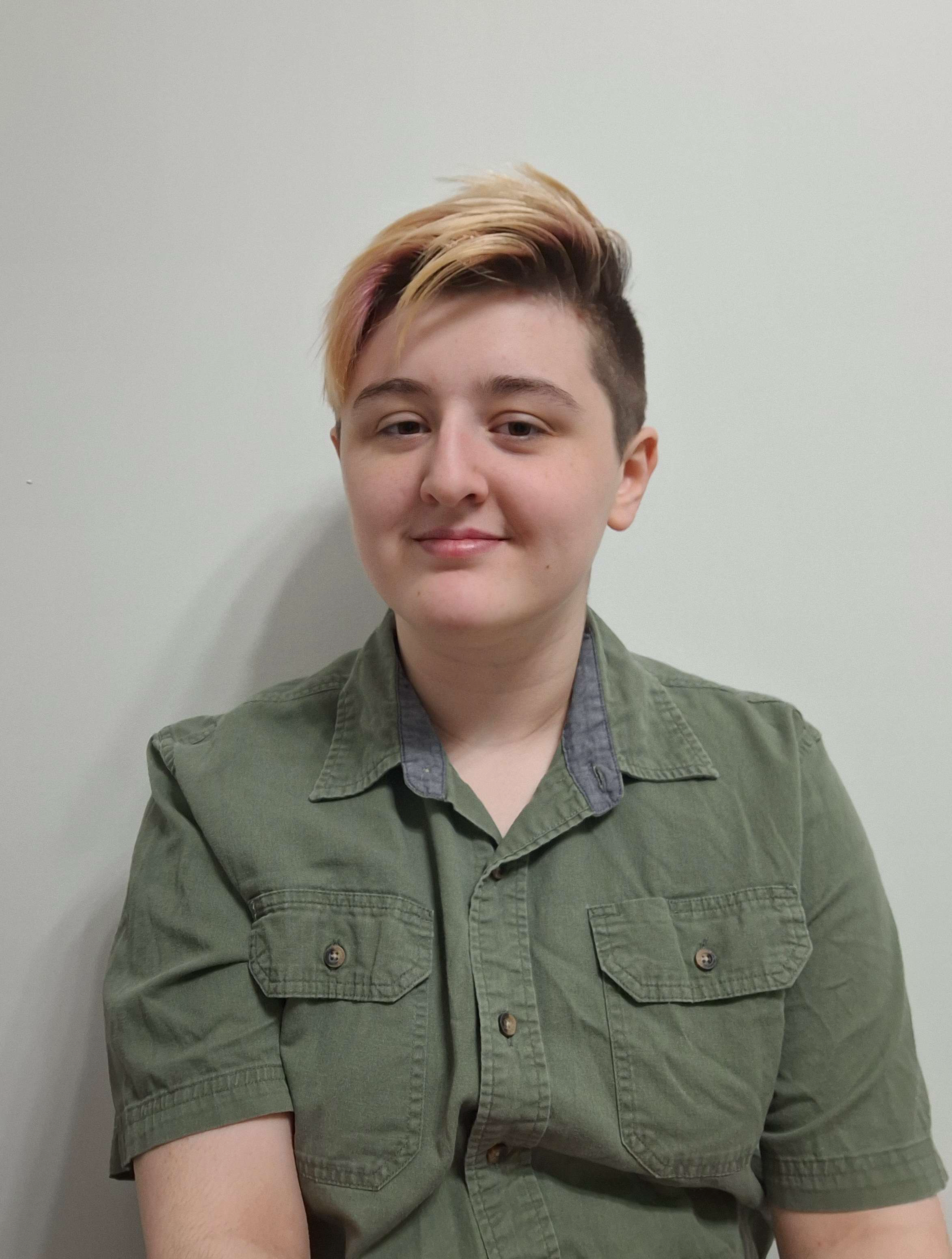 A person with short, styled hair that is blonde on top with dark brown sides wearing a green button-up shirt, standing against a plain light gray wall.