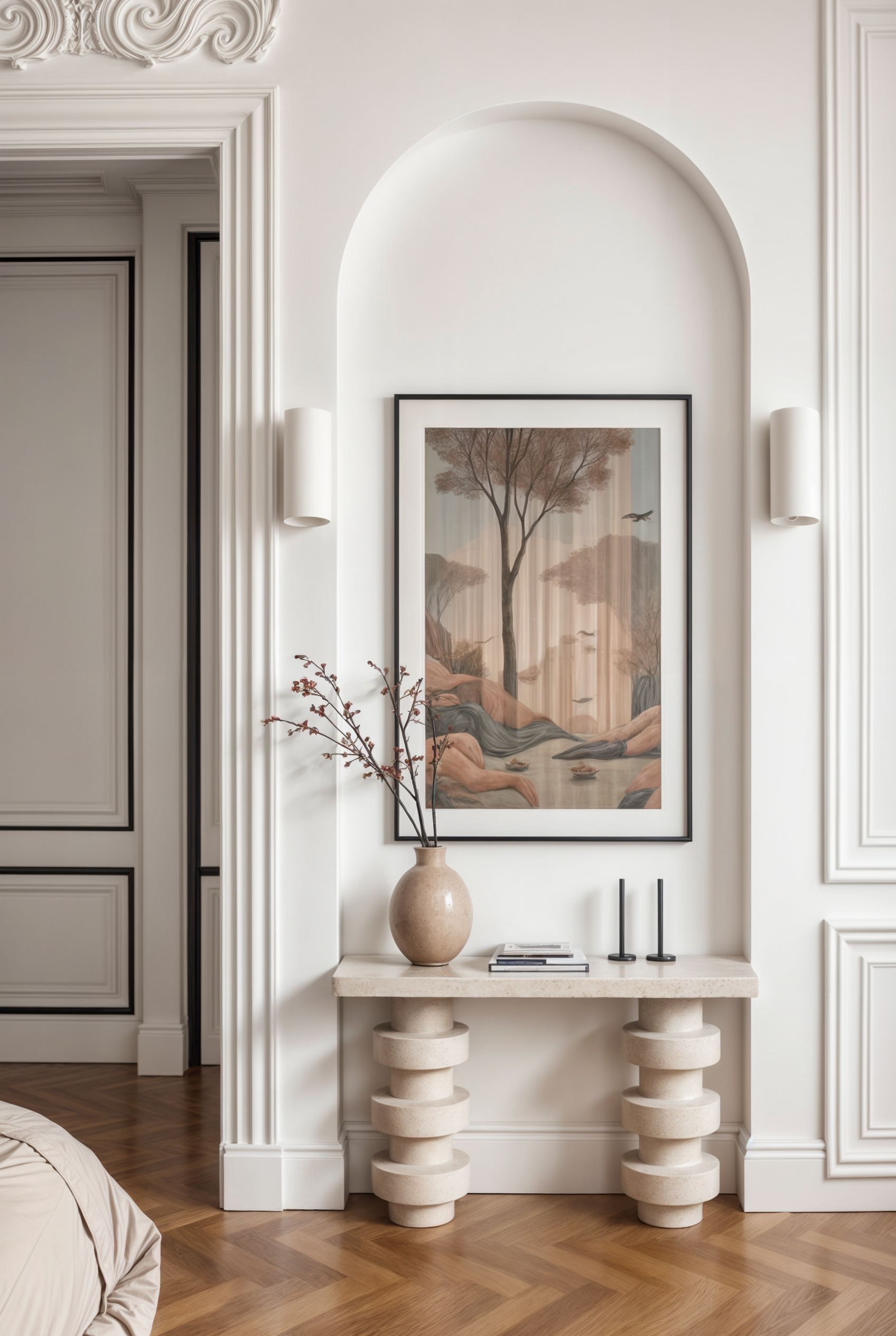 A beige console table with striped cylindrical legs, decorated with a beige vase holding a branch, two black candlesticks, and a framed painting of trees and mountains, set against white walls with decorative molding and two wall sconces.