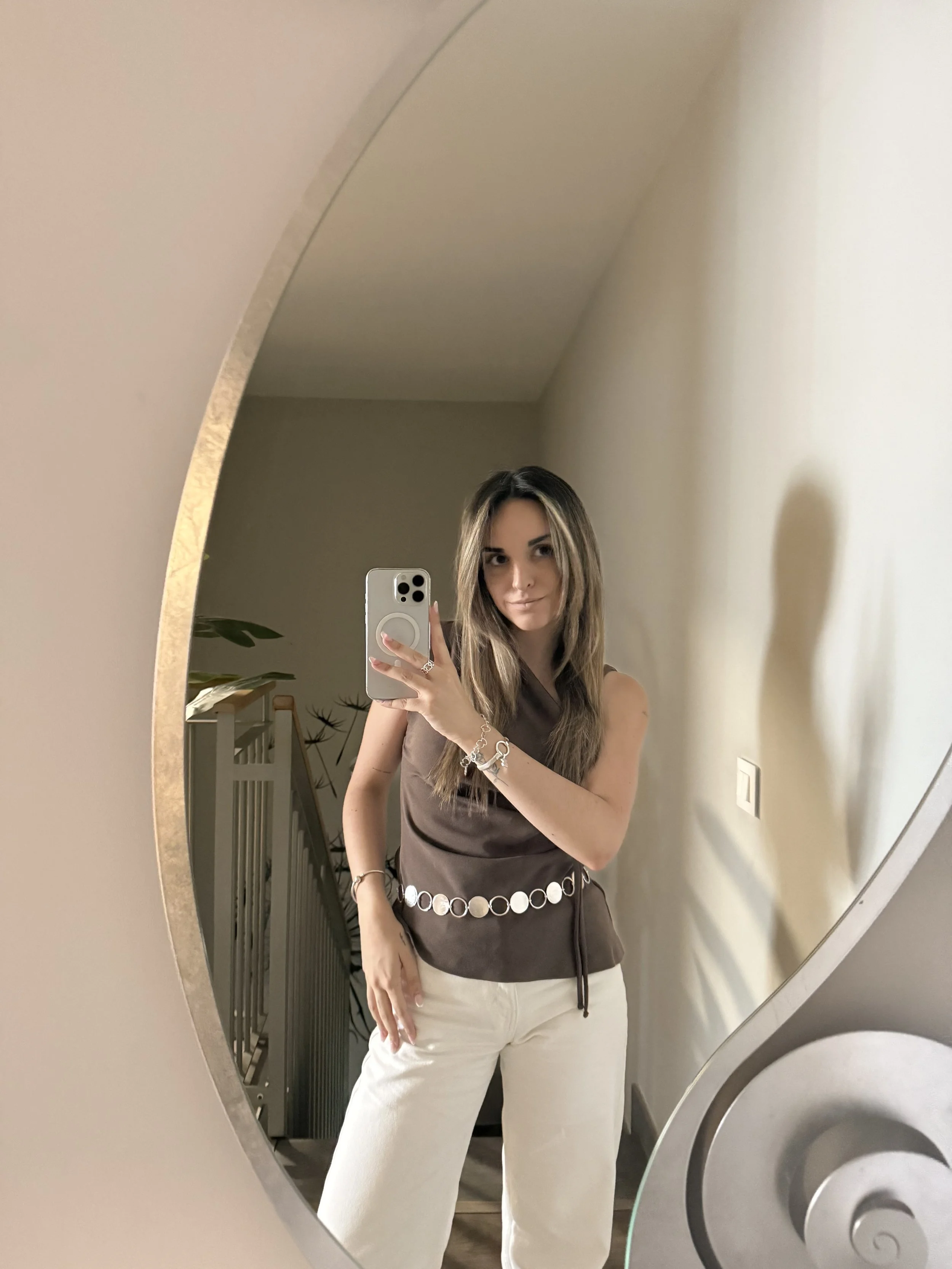 A woman taking a mirror selfie with her smartphone. She has long, wavy hair and is wearing a sleeveless brown top with decorative white circles on the hem and white pants. The background shows a staircase and a wall with some plants and decorative items.