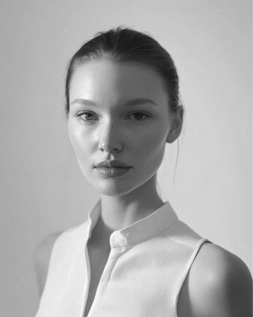 Black and white portrait of a young woman with her hair pulled back, wearing a sleeveless collared top, looking directly at the camera.