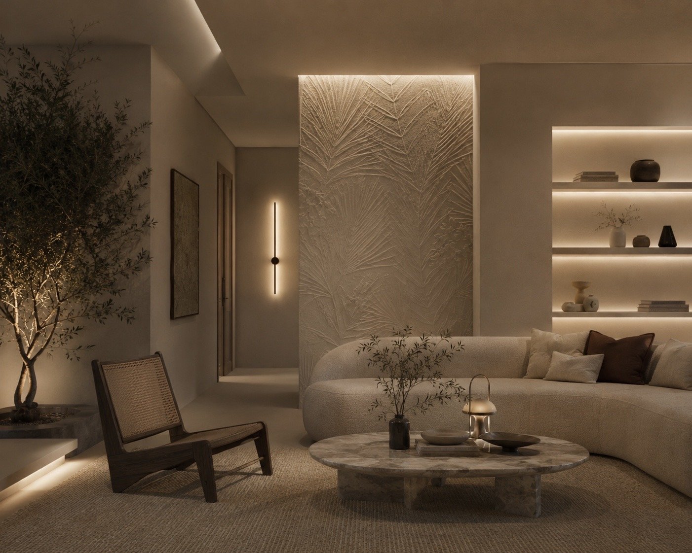 Living room with a beige curved sofa, wooden armchair, marble coffee table, decorative vases, a tree in a pot, wall-mounted light, and built-in shelves with decorative items, all illuminated with warm indirect lighting.