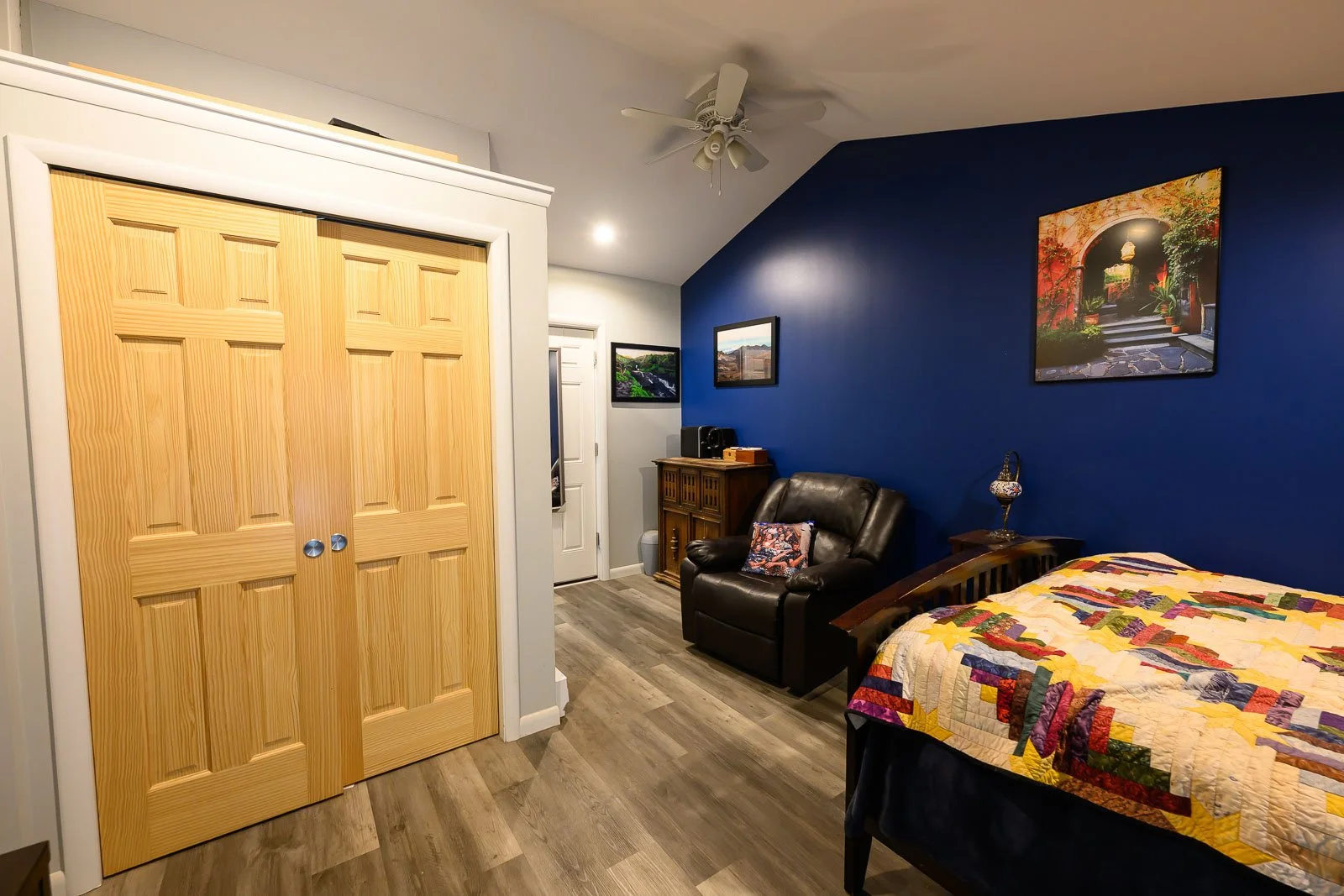 A bedroom with a blue accent wall, framed photographs, a black leather armchair, a wooden cabinet, a bed with a colorful quilt, a small side table with a decorative lamp, a ceiling fan, and a closet with wooden double doors.