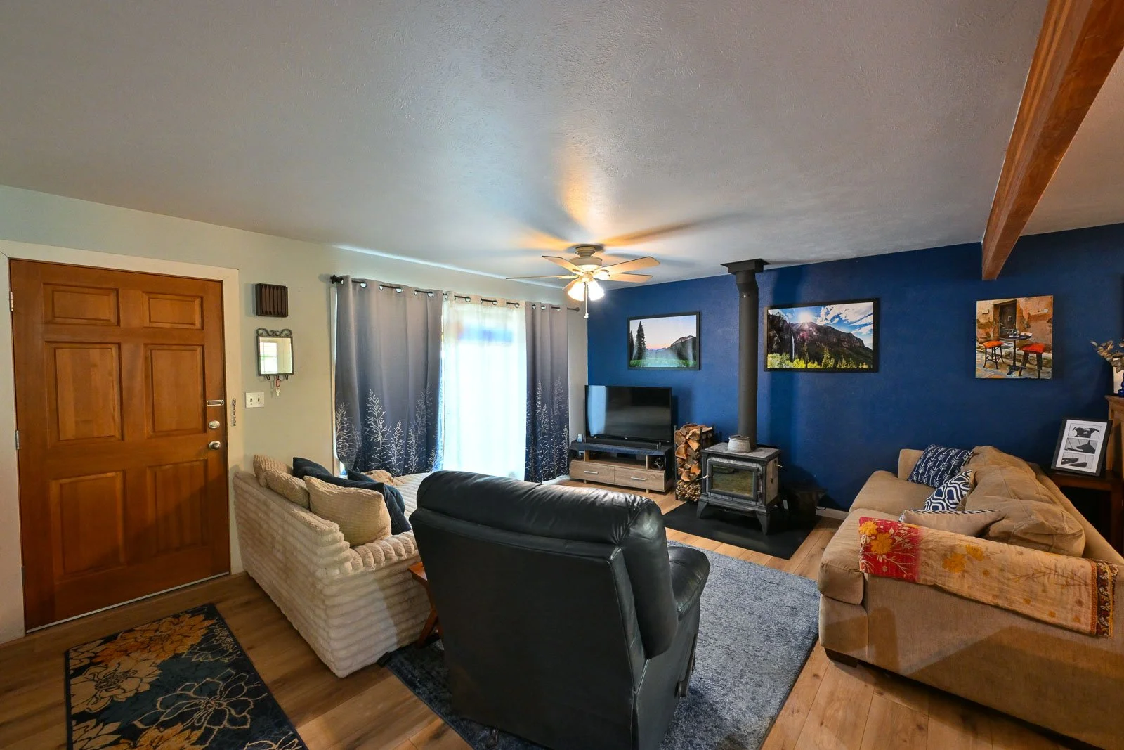 Living room with beige and black couches, a wooden door, a TV on a stand, a wood stove with stacked firewood, a dark blue accent wall, and framed landscape pictures, with curtains and ceiling fan.
