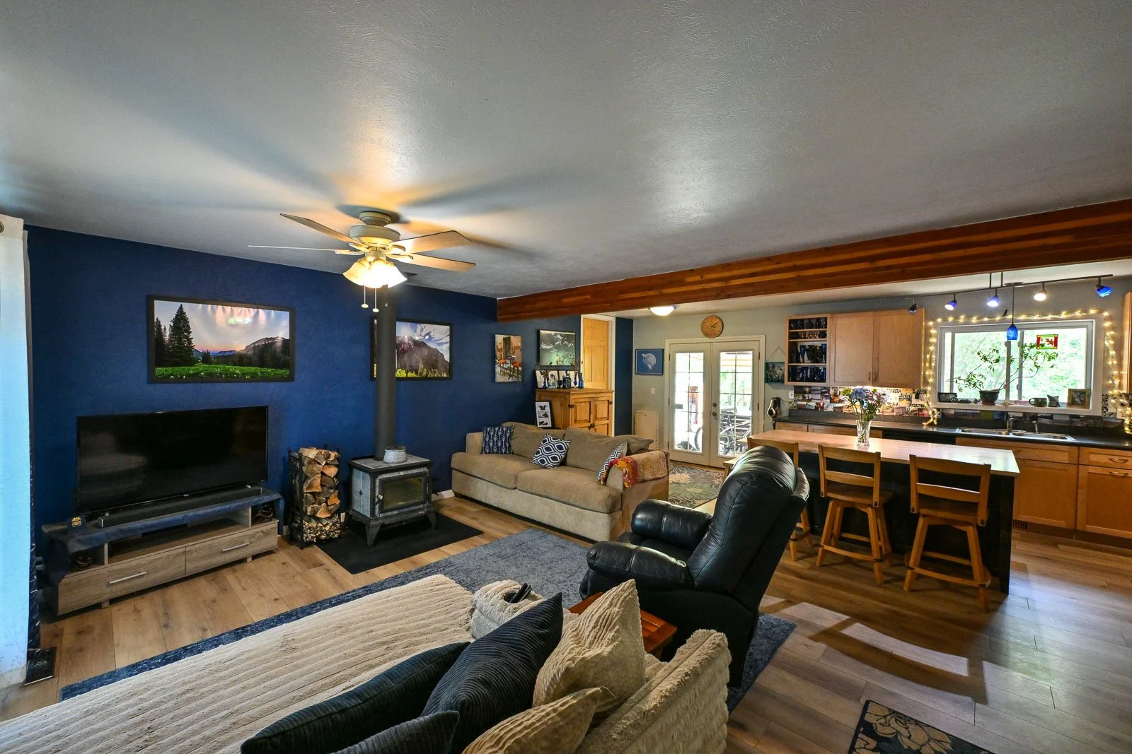 Living room with blue accent wall, TV, wood stove, beige and black leather furniture, and an open kitchen with wooden cabinets and a vase of flowers on the counter.