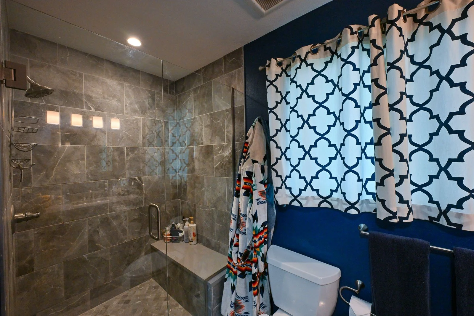 Bathroom with walk-in shower enclosed by glass, gray marble tiles on walls and floor, and a white toilet. A geometric patterned curtain covers the window, and towels hang on a bar.