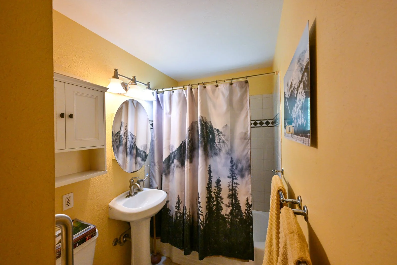 Bathroom with yellow walls, a white pedestal sink, an oval mirror, a shower with a mountain and trees printed shower curtain, yellow towels on a towel rack, a landscape photo on the wall, and a tile wall in the background.