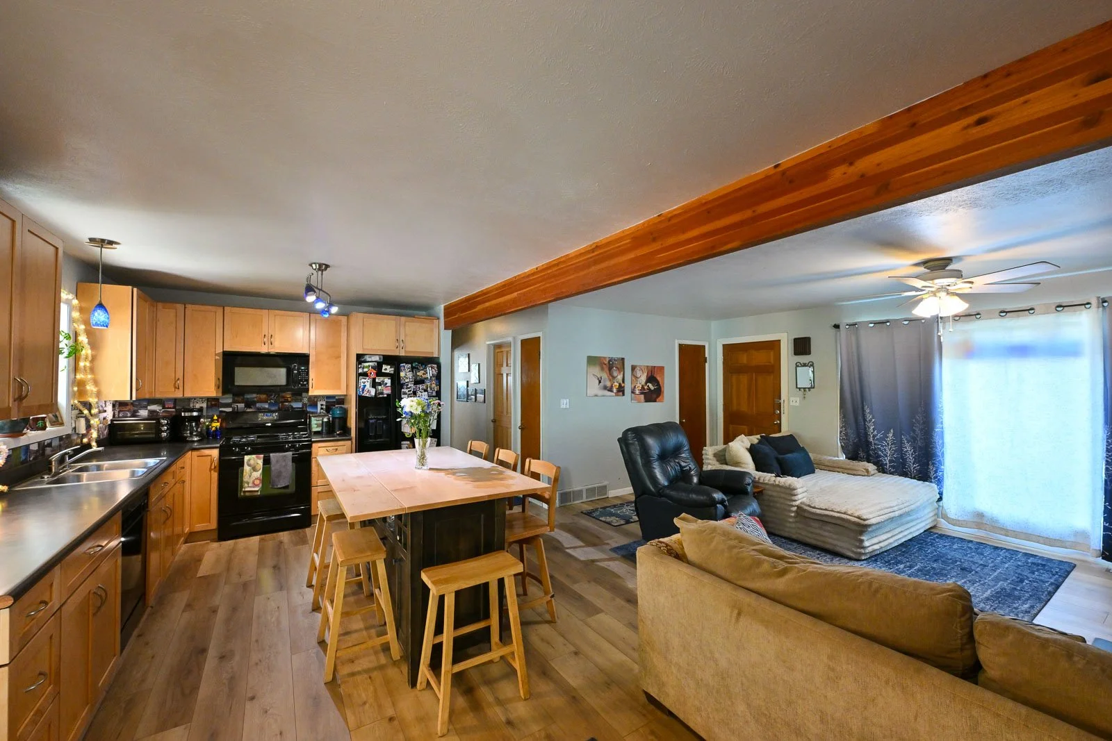 Open-concept living room and kitchen with wooden cabinets, black appliances, dining table with chairs, leather armchair, sofa, area rug, ceiling fan, and large window with curtains.