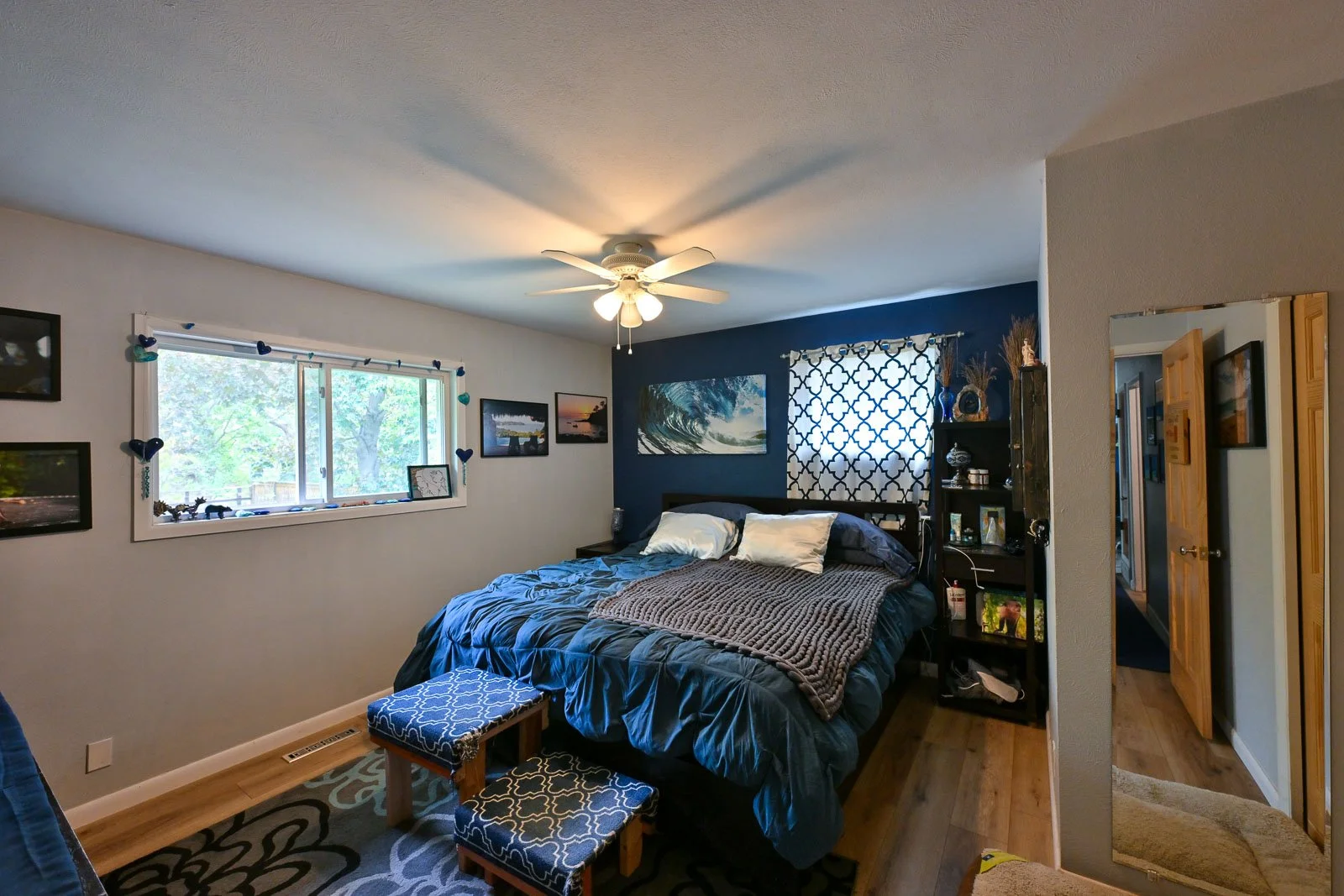 A cozy bedroom with a large window, a blue accent wall behind the bed, and a ceiling fan. The bed has white and dark blue pillows, a brown throw blanket, and a black headboard. There are framed pictures on the wall and a black bookshelf with decorati