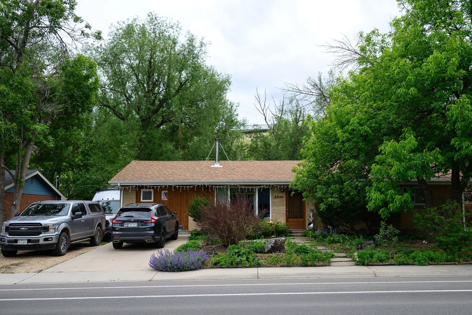 A house with a brown roof, brick and wood exterior, surrounded by green trees and a small garden in front. There are three vehicles parked in the driveway: a silver truck, a black SUV, and a white vehicle. The house has a front porch, and string ligh