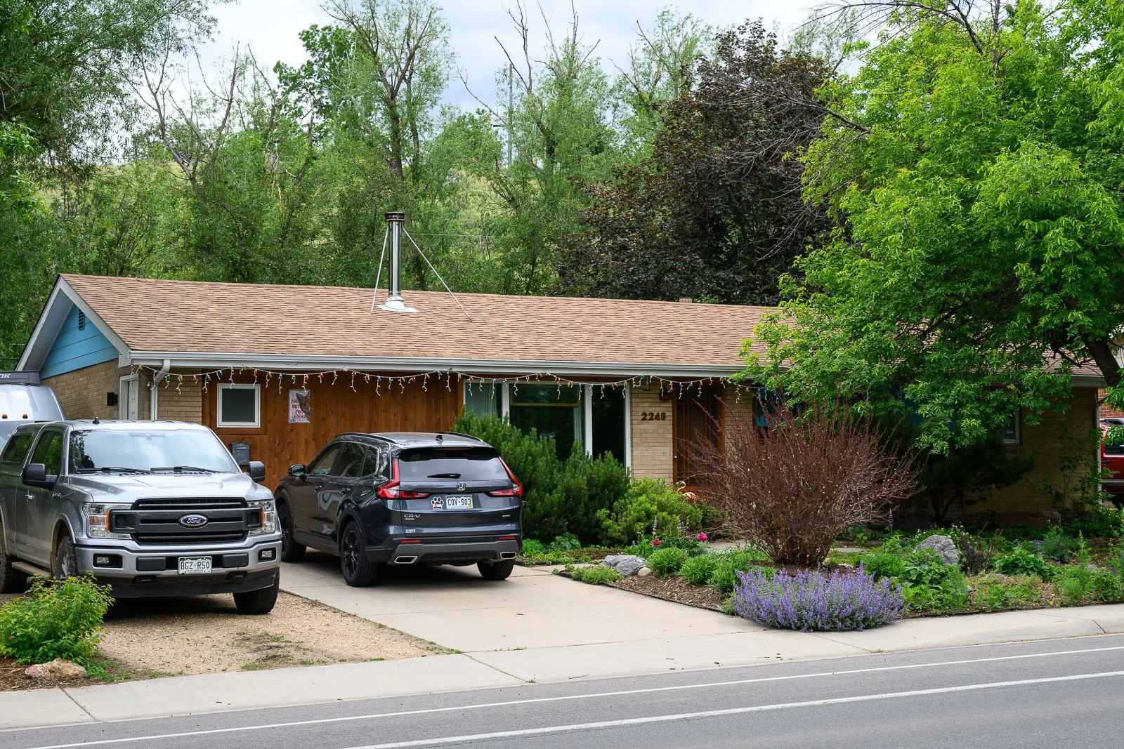 A single-story house with a brown brick exterior and a brown shingle roof. The house is decorated with string lights. There is a garden with colorful flowers and bushes in front of the house, and a large green tree to the right. Several cars are parked in the driveway, including a silver pickup truck and a black SUV. The house number, 2240, is visible near the front door.