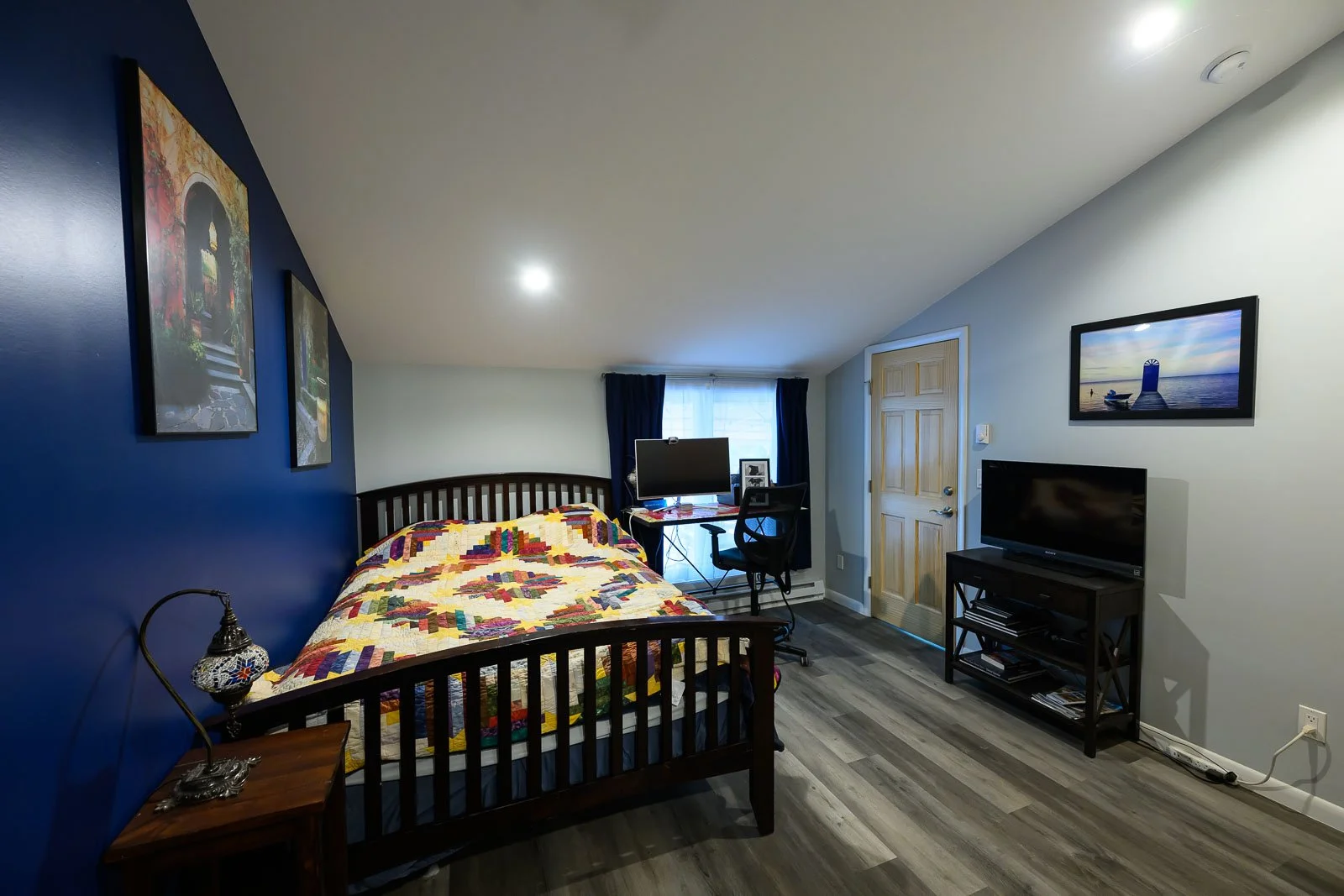 A cozy bedroom with a bed covered in a colorful quilt, a nightstand with a lamp, a small desk with a computer monitor, and a flat-screen TV on a black stand. The room has a blue accent wall, framed artwork, a window with dark curtains, and wood floor