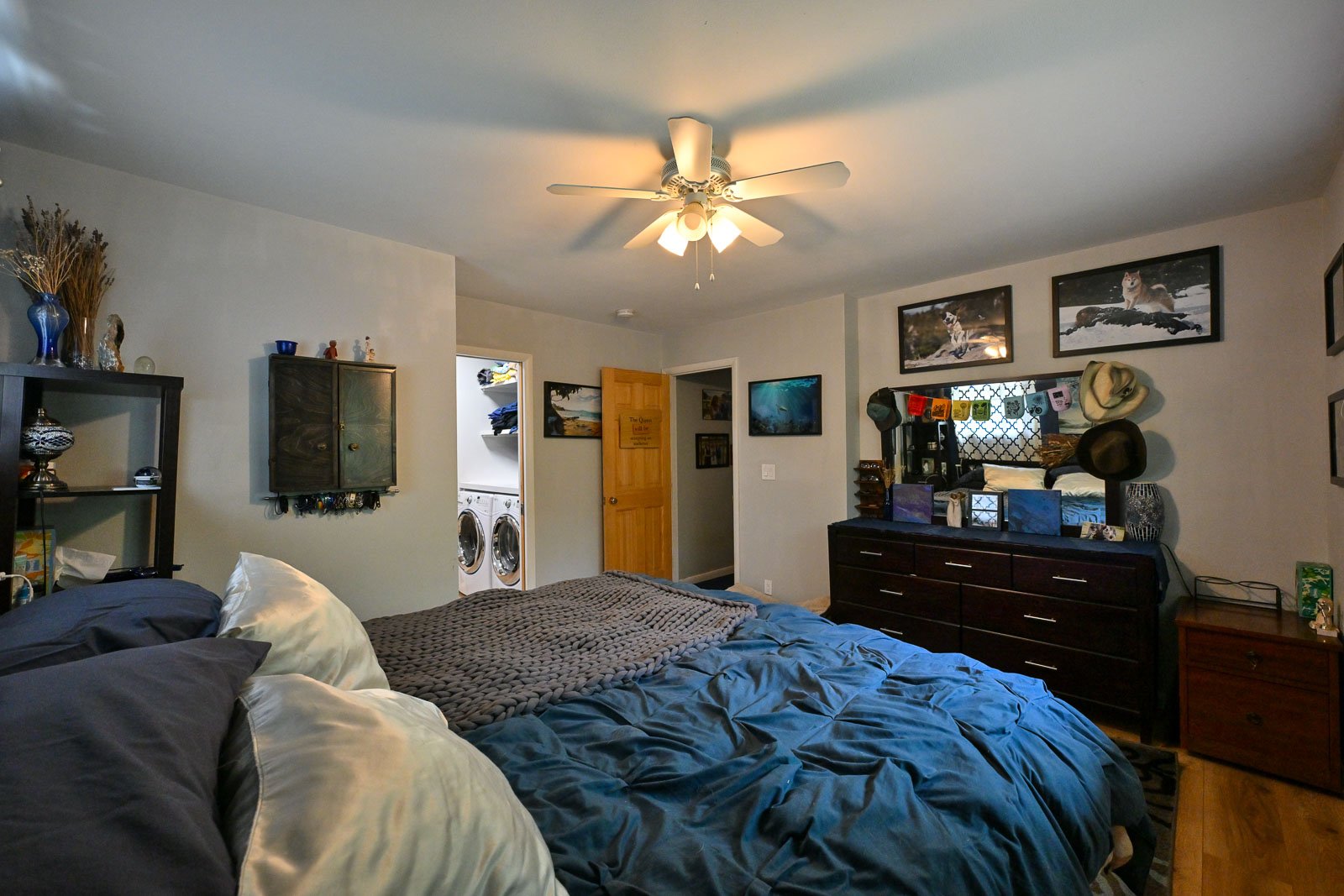 A bedroom with a bed featuring a dark gray blanket and pillows, a ceiling fan, a dresser with a mirror, and photographs of dogs on the wall. An open door reveals a laundry area with a washer and dryer.