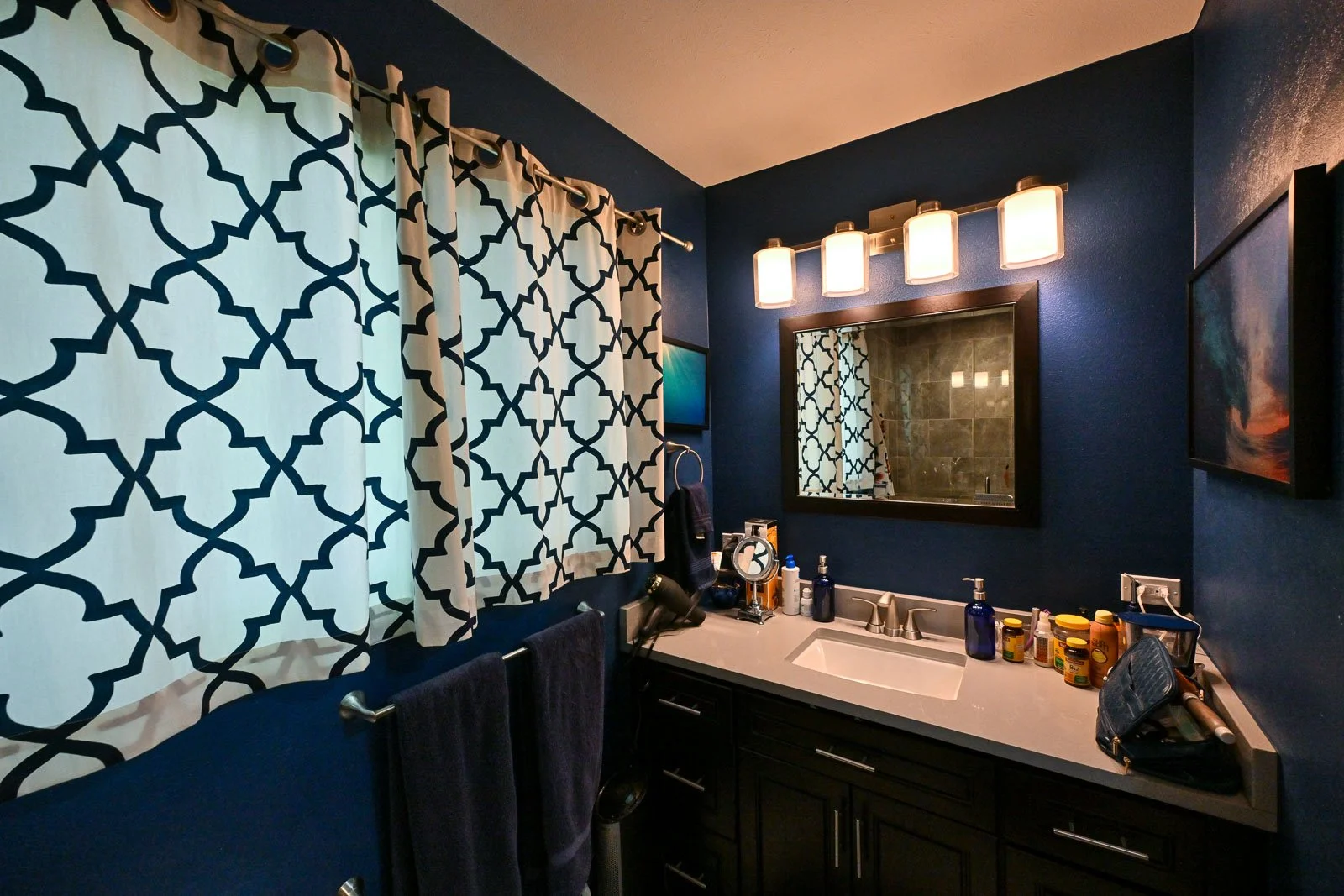 Bathroom with navy blue walls, patterned curtains, rectangular mirror above a white sink, modern light fixture, various bottles and toiletries on the countertop, and a wall-mounted TV.