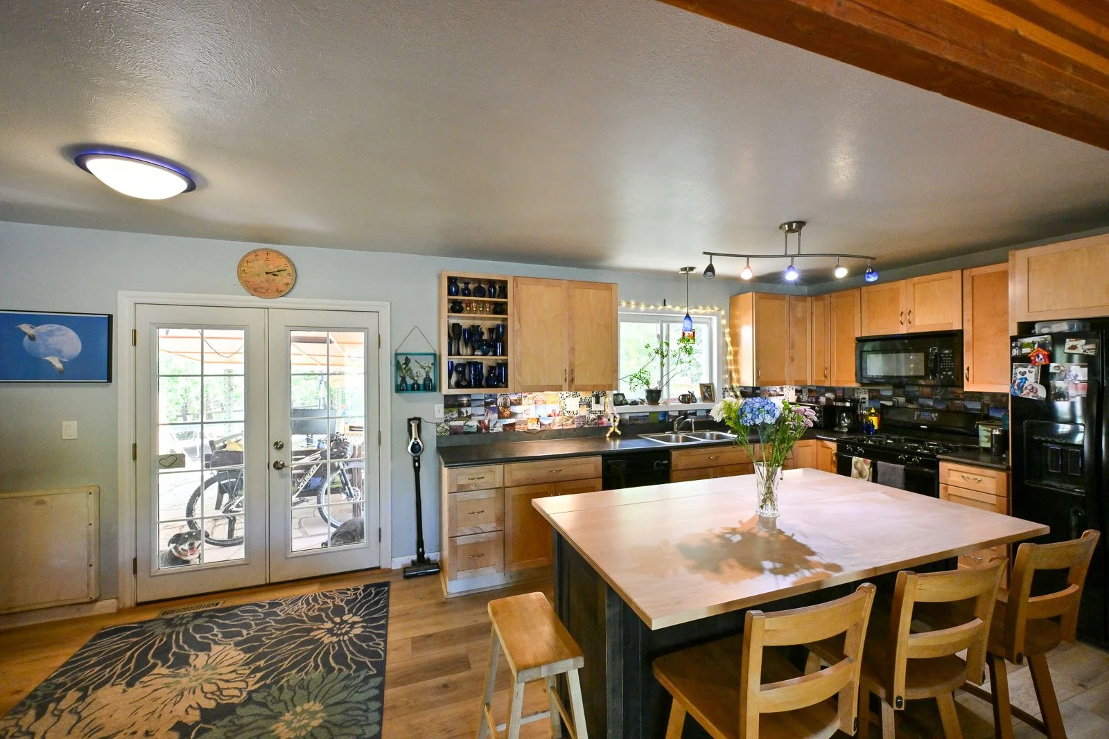 A kitchen with wooden cabinets, black appliances, and a large wooden dining table with chairs. French doors lead outside, with a view of bicycles on a porch. The kitchen has a colorful tile backsplash and a vase of flowers on the table.