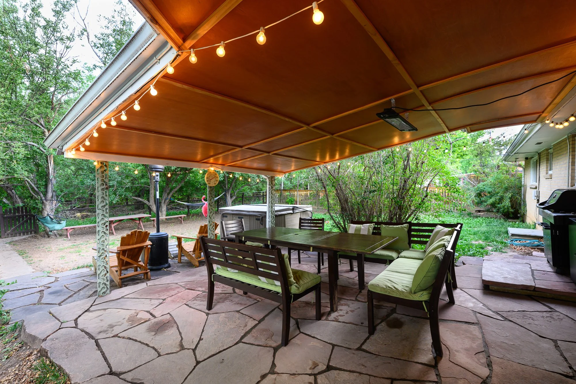 Outdoor covered patio area with string lights, a dining table with chairs, surrounded by trees and greenery in a backyard.