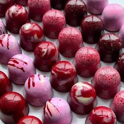 Assorted glossy chocolate truffles in shades of pink, red, and purple, some with drizzles or speckles, arranged in rows.
