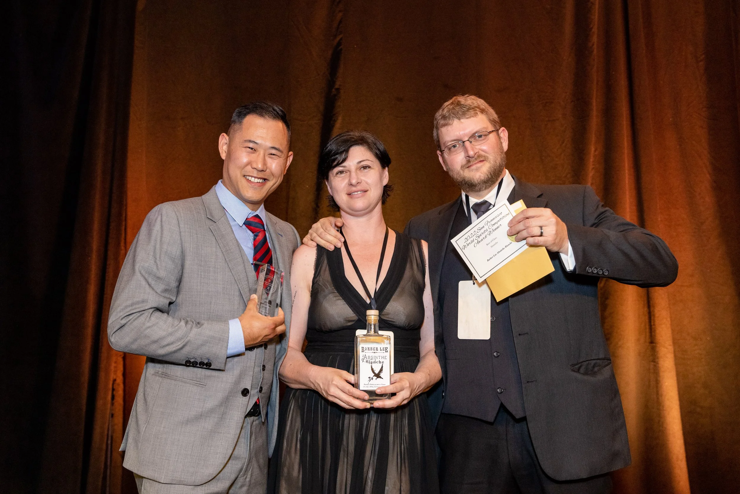 Three individuals standing together at an award ceremony. The man on the left is wearing a grey suit and a striped tie, smiling and holding a clear glass award. The woman in the middle has short dark hair, wearing a black dress, and holding a bottle of absinthe. The man on the right has glasses, a beard, and is wearing a black tuxedo, holding a certificate and pointing to it. They are in front of a brown curtain.