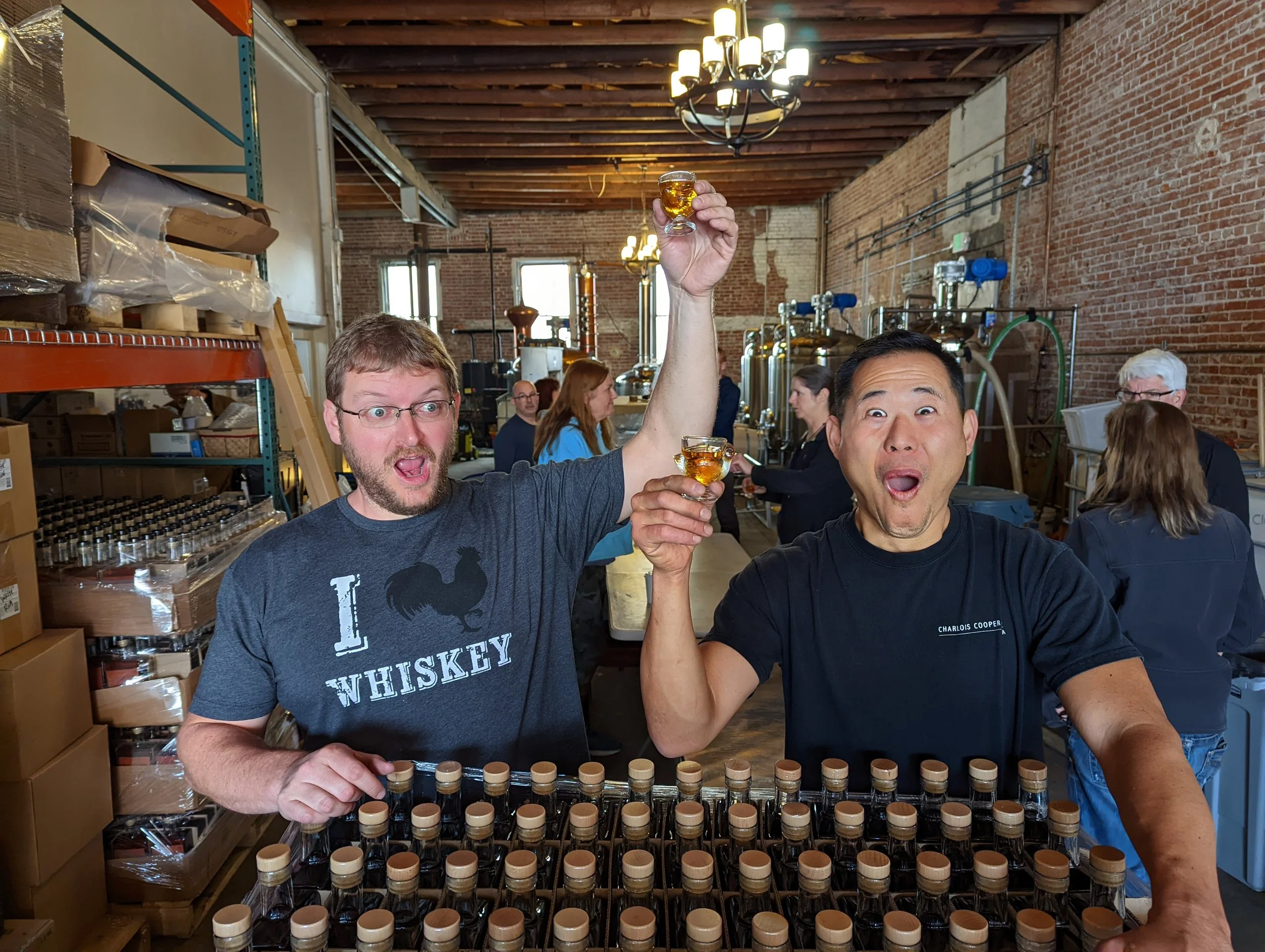 Two men smiling and celebrating with glasses of whiskey in a brewery with brick walls and brewery equipment in the background.