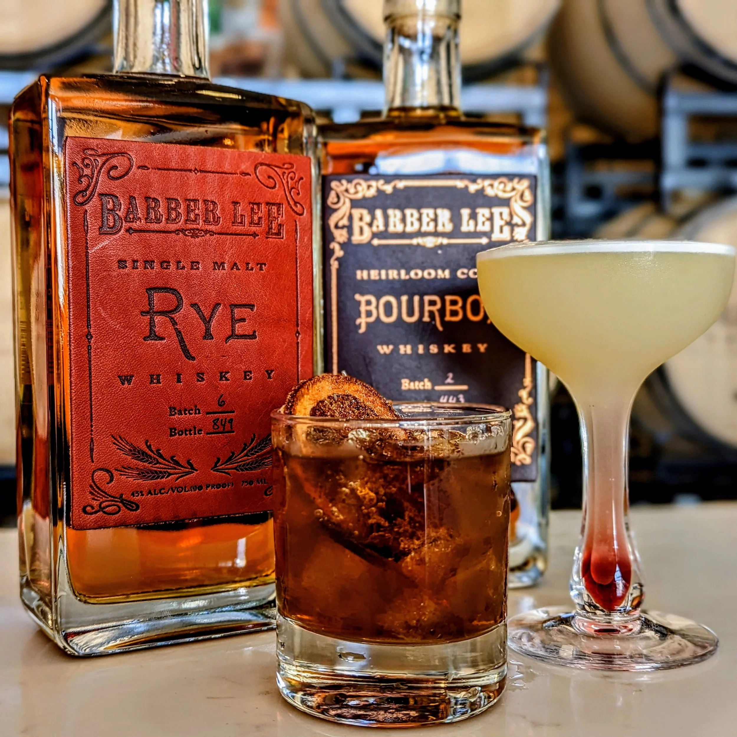 Close-up of whiskey bottles labeled 'Barber Lee Single Malt' and 'Heirloom Coffee Bourbon,' with glasses of whiskey on a white surface.