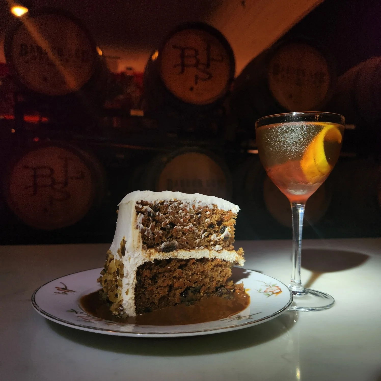 A slice of chocolate cake with icing on top, served on a floral plate, next to a glass of sparkling water with lemon, with wine barrels in the background.