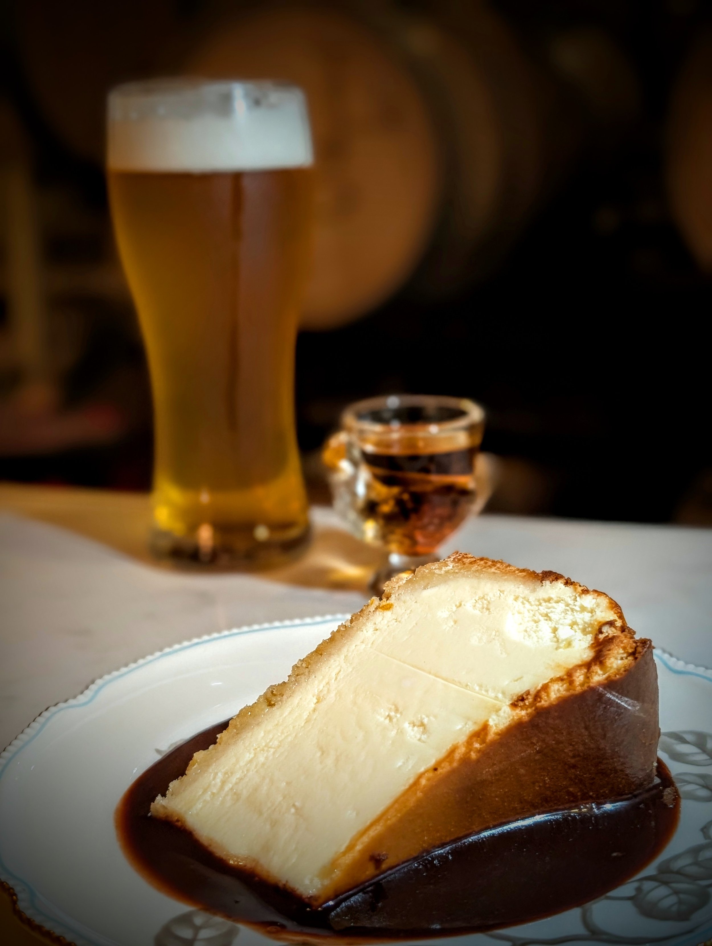 A slice of cheesecake with a golden-brown crust on a white plate, topped with chocolate sauce. In the background, there is a tall glass of beer with foam on top, and a small shot glass filled with a dark liquid.