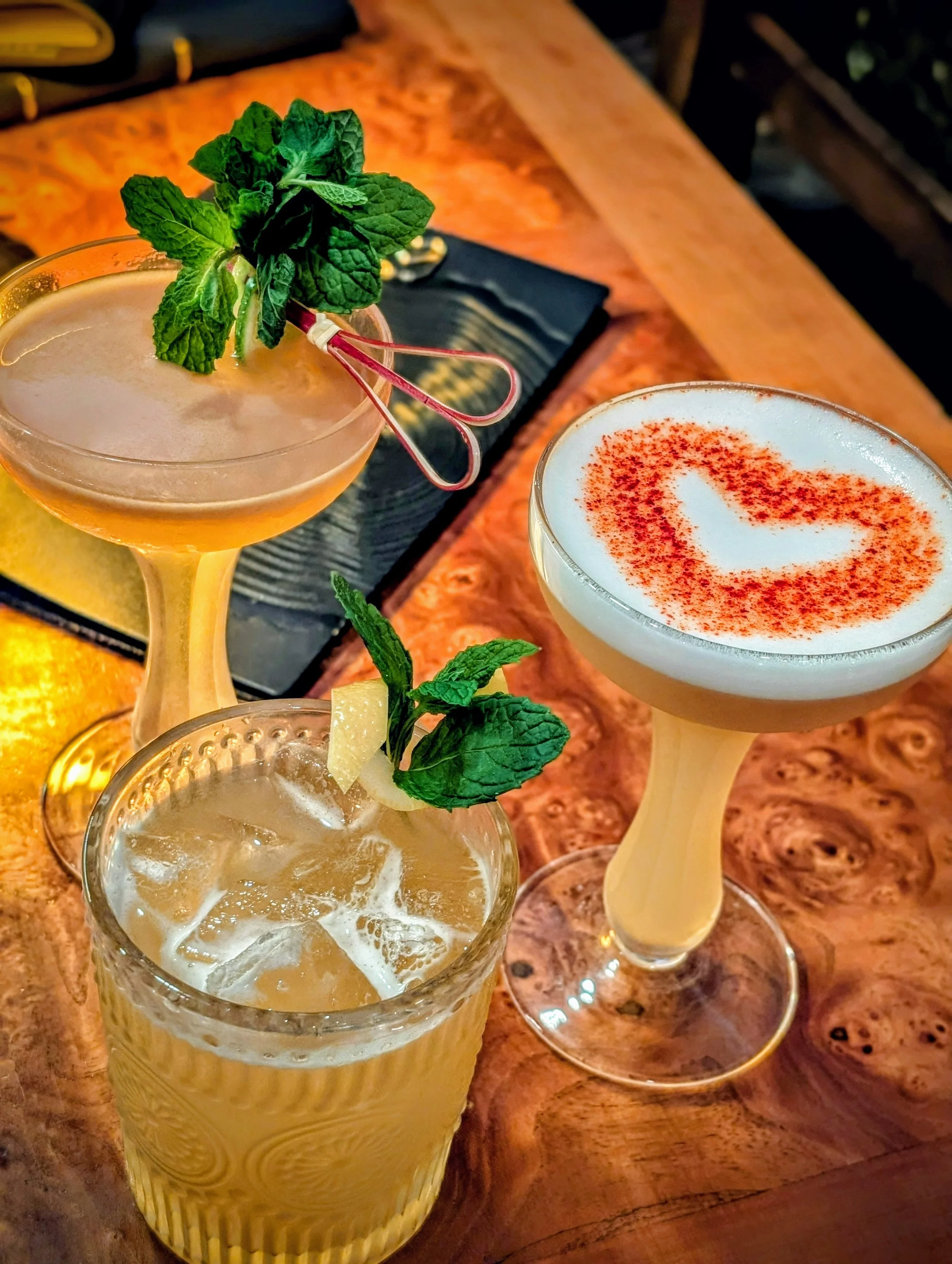 Three cocktails on a wooden table. The first cocktail is in a tall, textured yellow glass, garnished with a sprig of mint and a lemon slice. The second cocktail is in a coupe glass, topped with foam and sprinkled with red spice, garnished with mint and a lemon wedge. The third cocktail is in a short glass, filled with ice and garnished with mint and a lemon wedge.