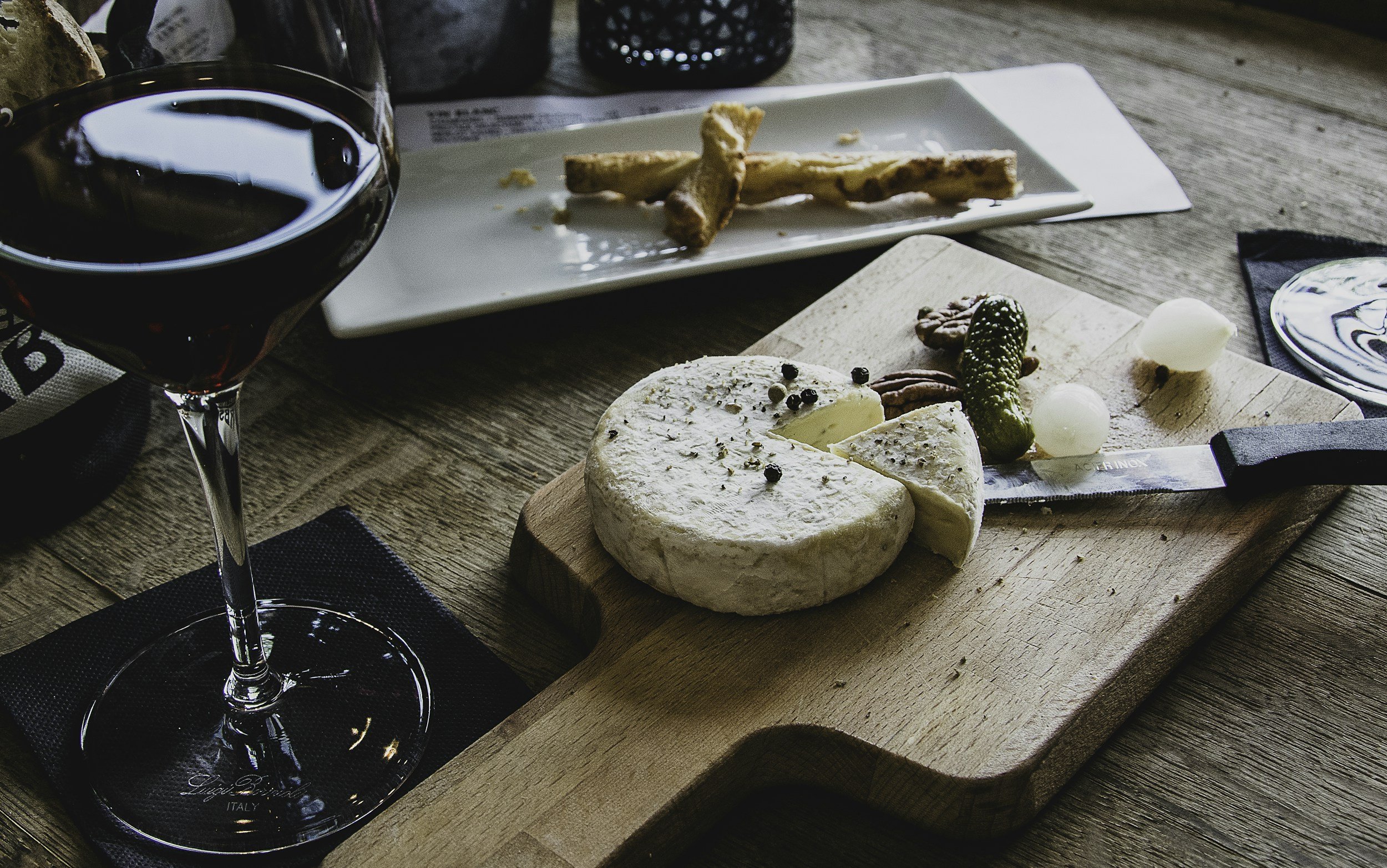 A glass of red wine and a cheese platter with a wheel of cheese, pickles, and nuts on a wooden serving board, set on a rustic wooden table.