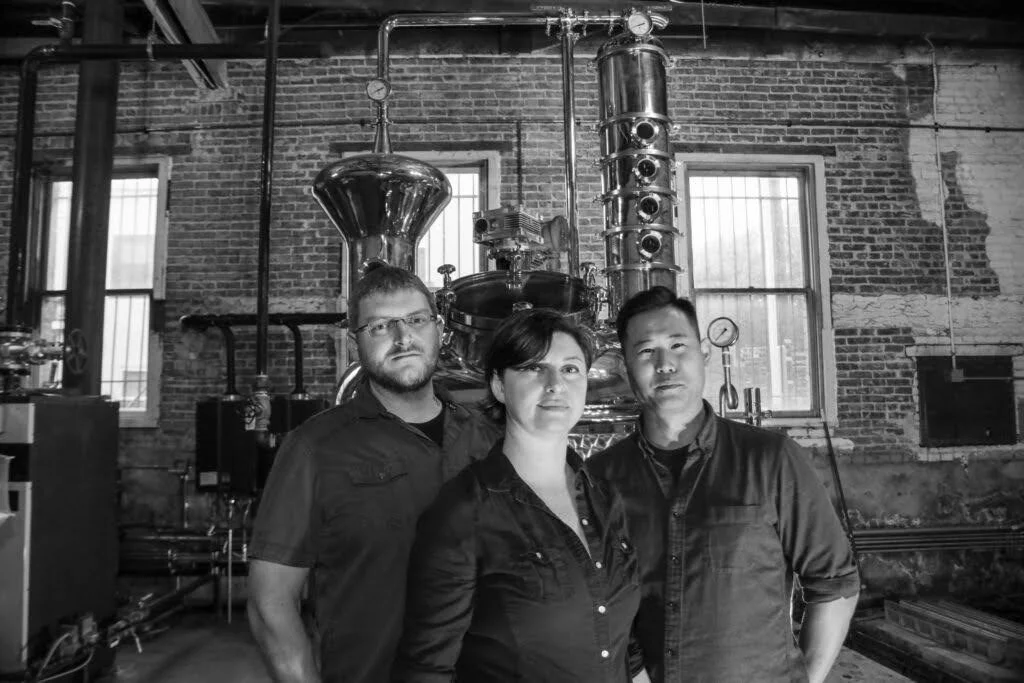 Three people standing in front of distillation equipment in an industrial brewery setting.
