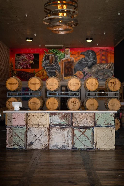 Barrels with the logo 'Barbers Lee' arranged on a counter in front of a colorful mural featuring various animals and a cityscape. The mural includes a black bird, a snake, an owl, and other creatures, and the setting appears to be a bar or salon with a rustic, industrial design.