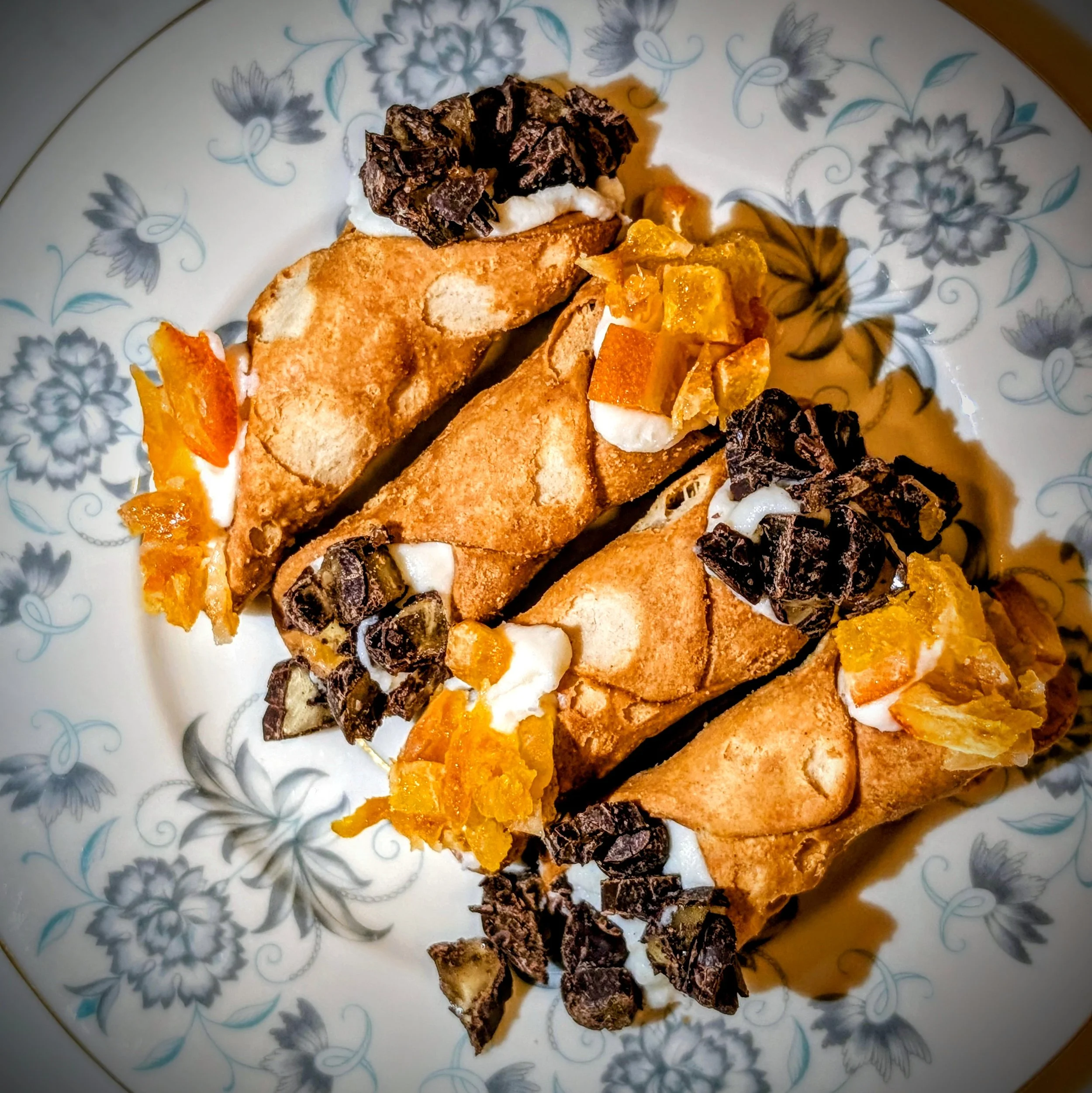 A decorative plate with a floral pattern holds a partially eaten slice of cake. The cake has a vanilla or light-colored base with orange and yellow fruit pieces, topped with whipped cream and a chocolate garnish. There are additional fruit pieces arranged around the cake on the plate.