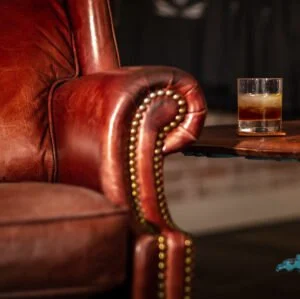 Close-up of a brown leather armchair with decorative brass nailhead trim, next to a wooden table with a glass of layered drink.