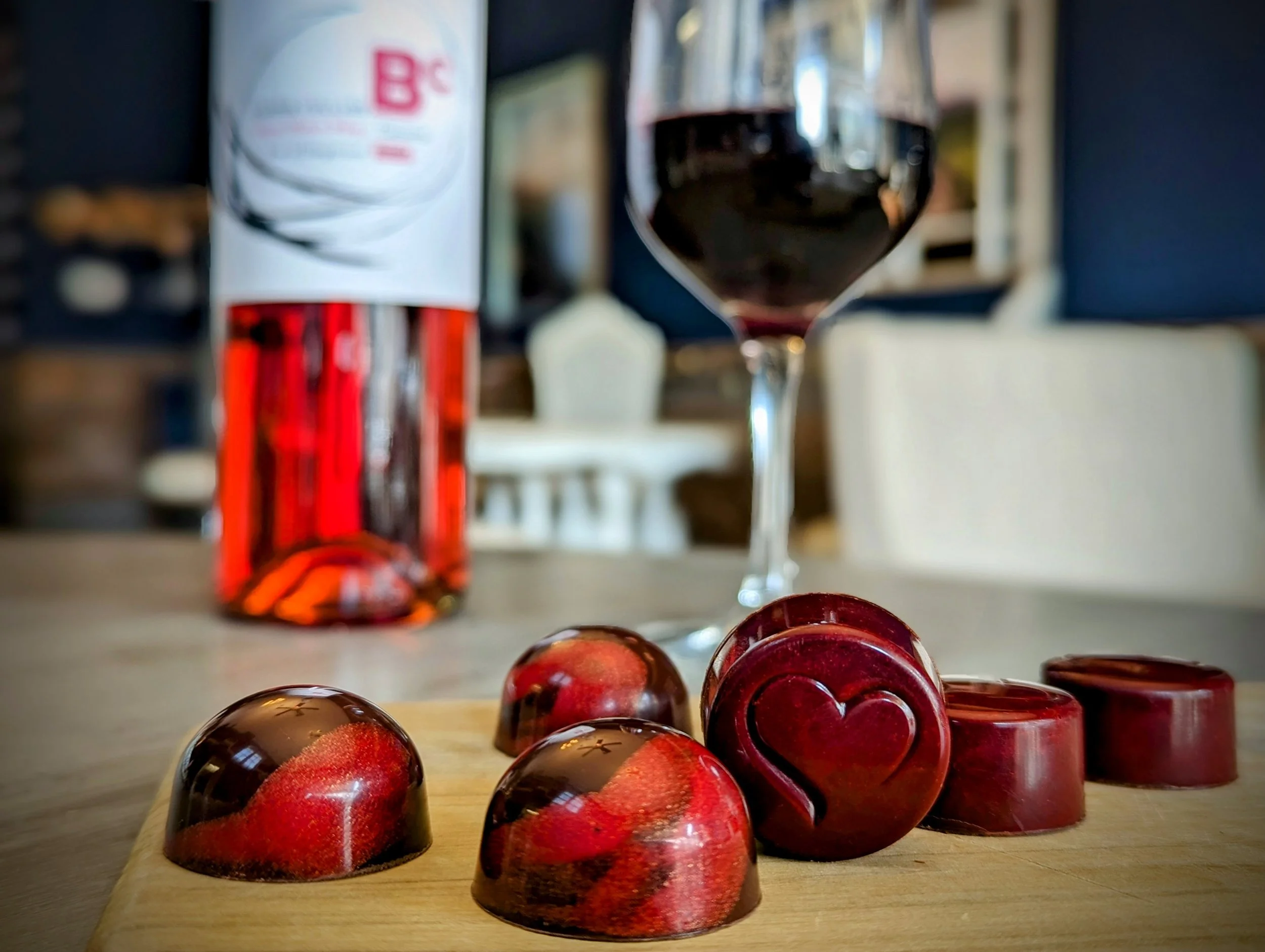A collection of red and black marbled chocolates on a light wooden surface with a glass of red wine and a white container in the blurred background.
