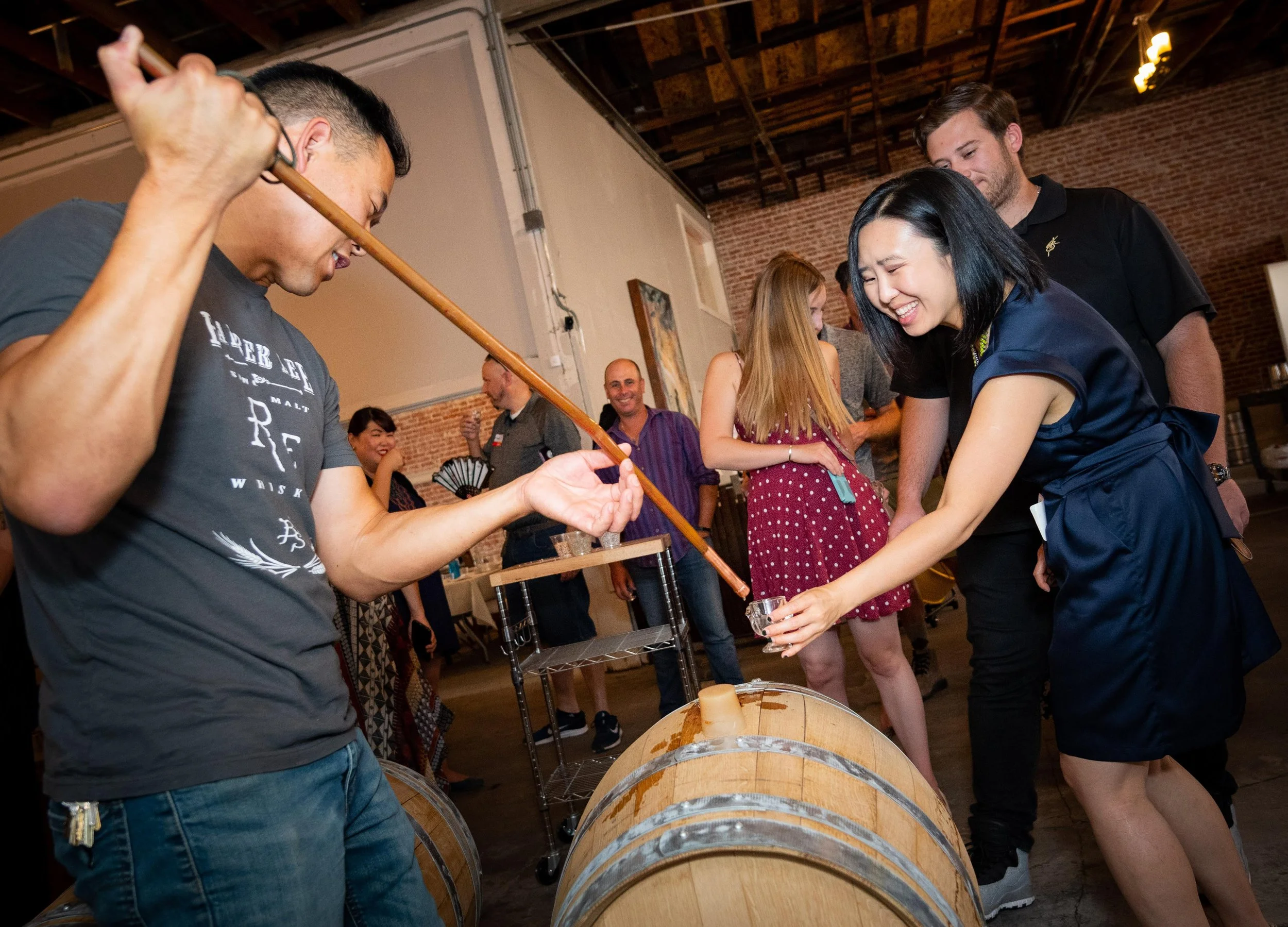 People participating in a wine or drink tasting event indoors, with a person pouring a liquid into a glass from a barrel, and others smiling and enjoying the activity.