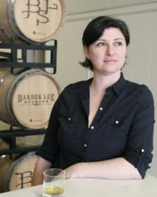 A woman with short dark hair wearing a dark shirt sitting in front of barrels at a winery or brewery, with a glass of white wine on the table.