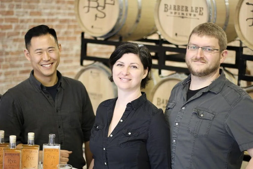 Three people standing in front of barrels with labeling, smiling, with bottles of liquor in the foreground.