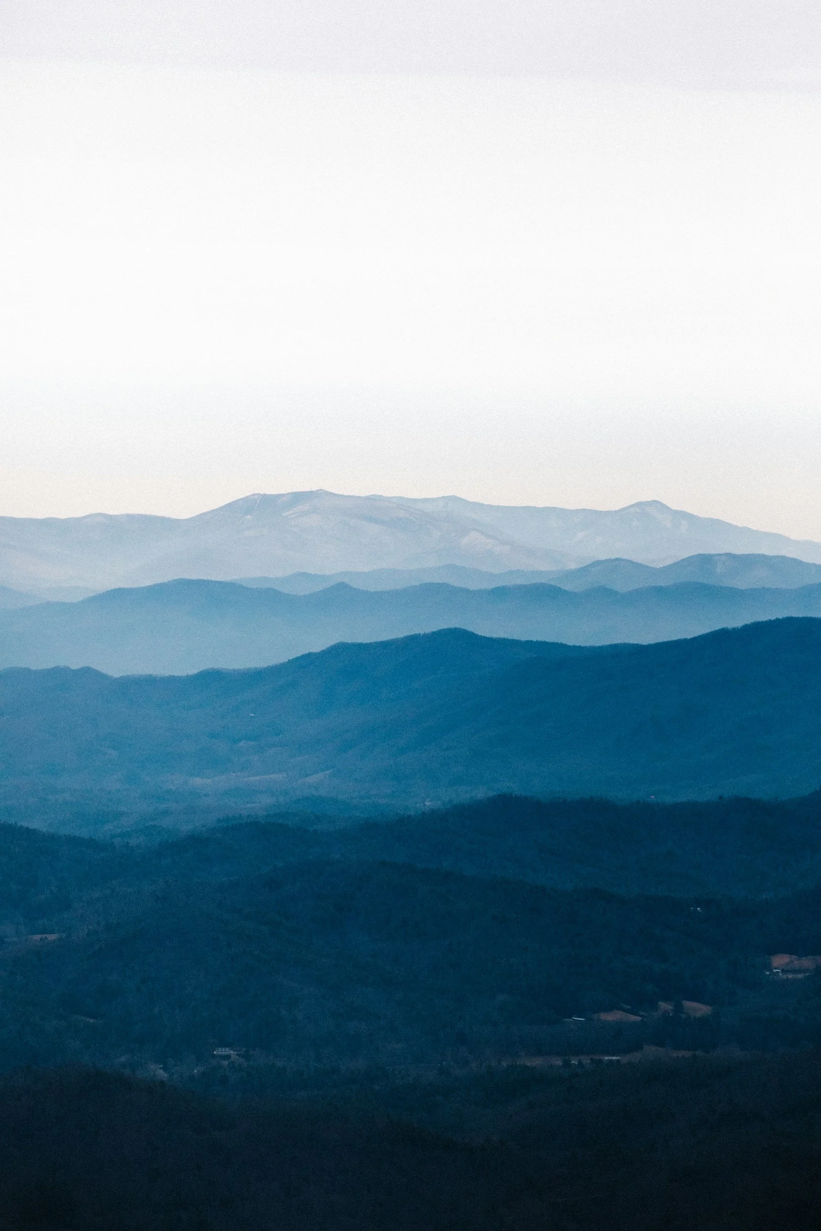 Layered mountain ranges with various shades of blue and green under an overcast sky.