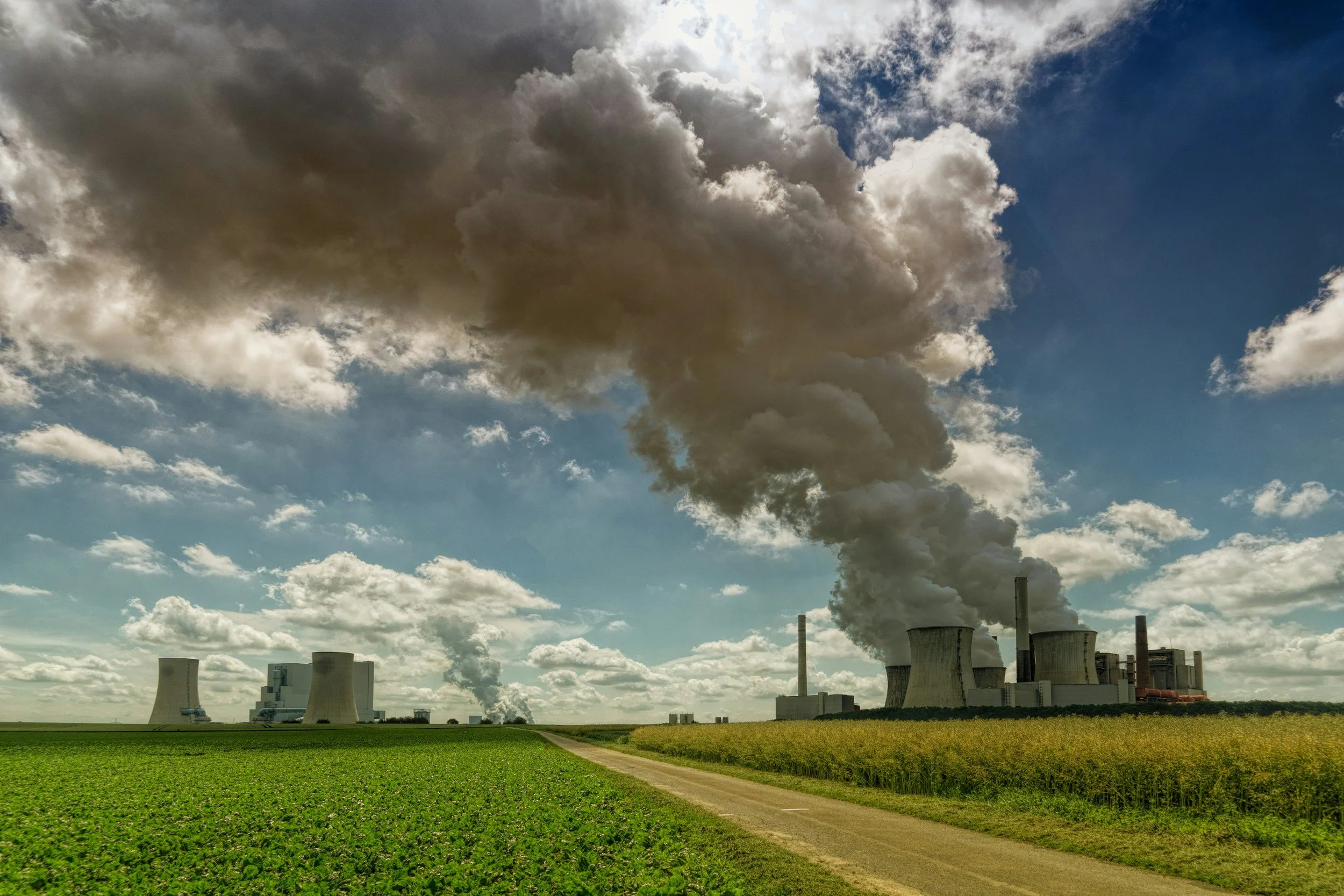 A power plant with multiple cooling towers emitting thick smoke into the sky, set against a partly cloudy sky with sunlight breaking through.