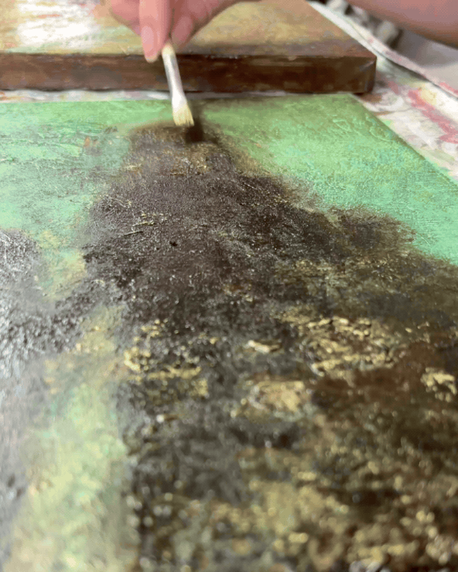 Close-up of a person using a brush to apply gold leaf or metallic gold dust onto a textured, painted surface, possibly for an art project or restoration.