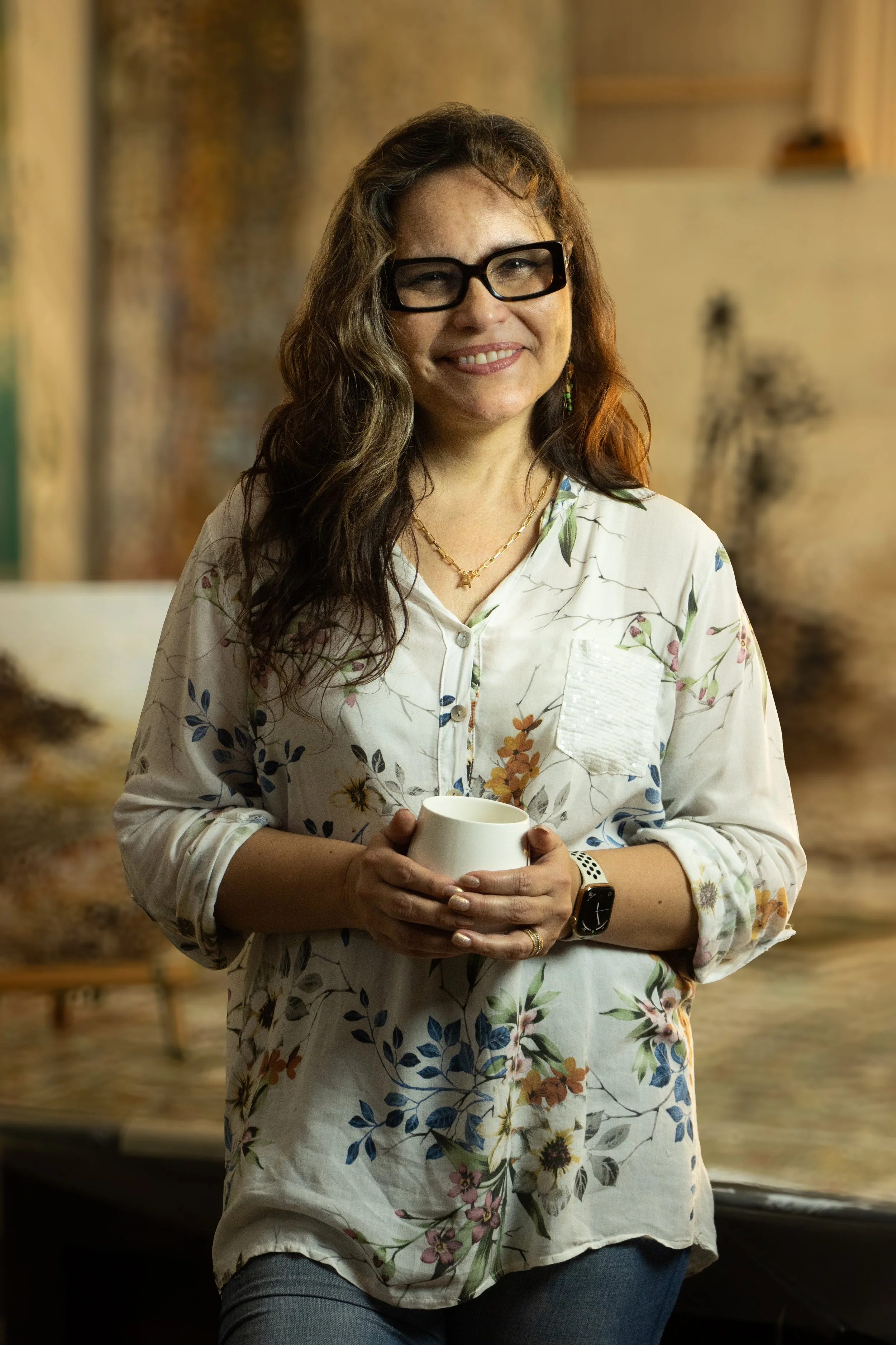 A woman with long wavy brown hair, wearing black glasses, a floral blouse, and a smartwatch, holding a white mug, standing indoors with a blurred background.