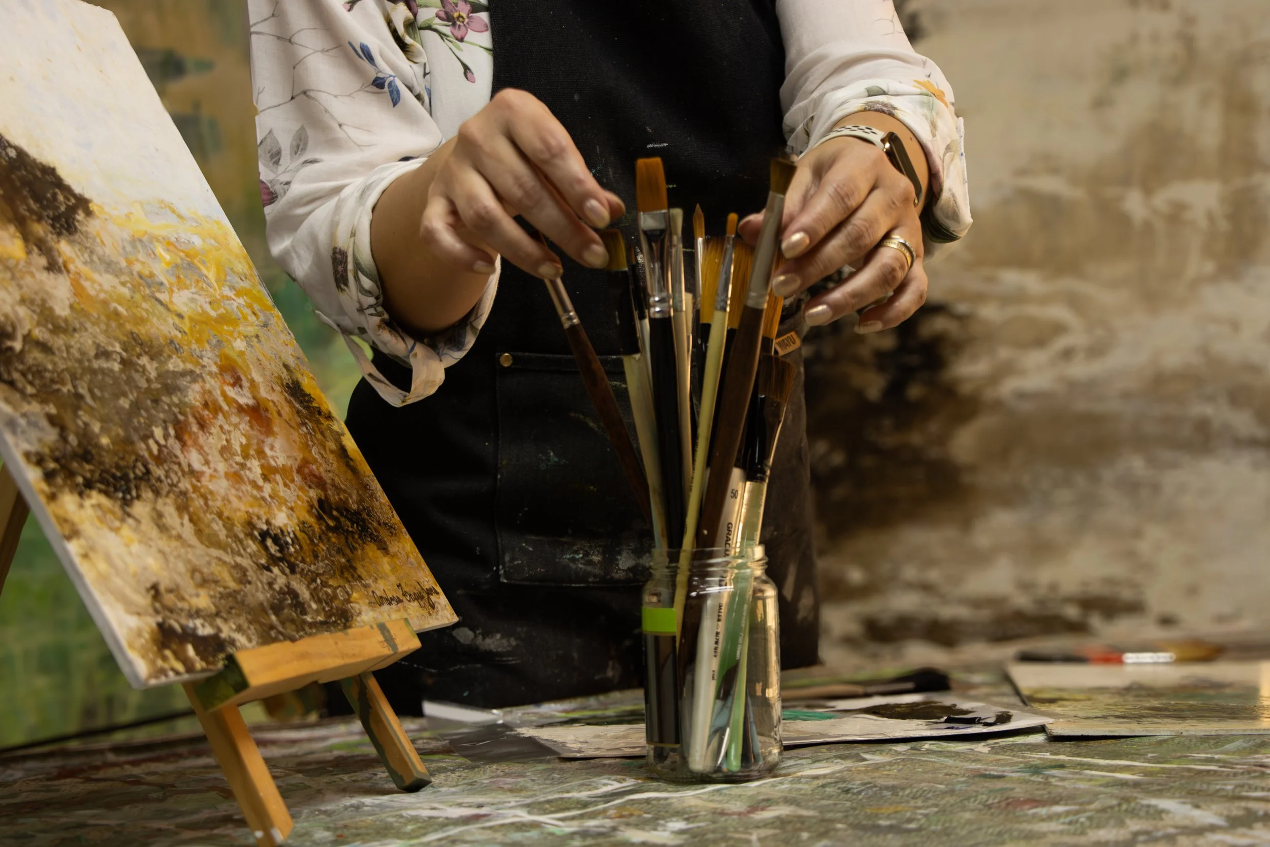 Close-up of hands of an artist holding paintbrushes in a glass jar, with an abstract painting on an easel and a textured brown wall in the background.
